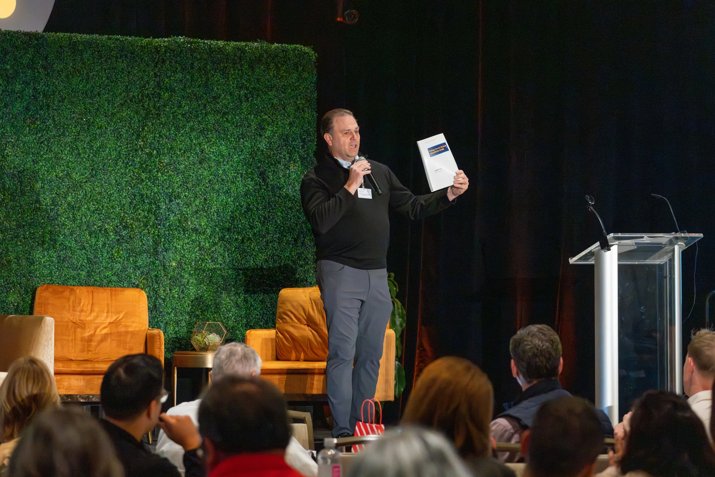 A man holding a microphone and a booklet, speaking on stage at a conference with an audience watching. There are orange chairs and a green wall backdrop.