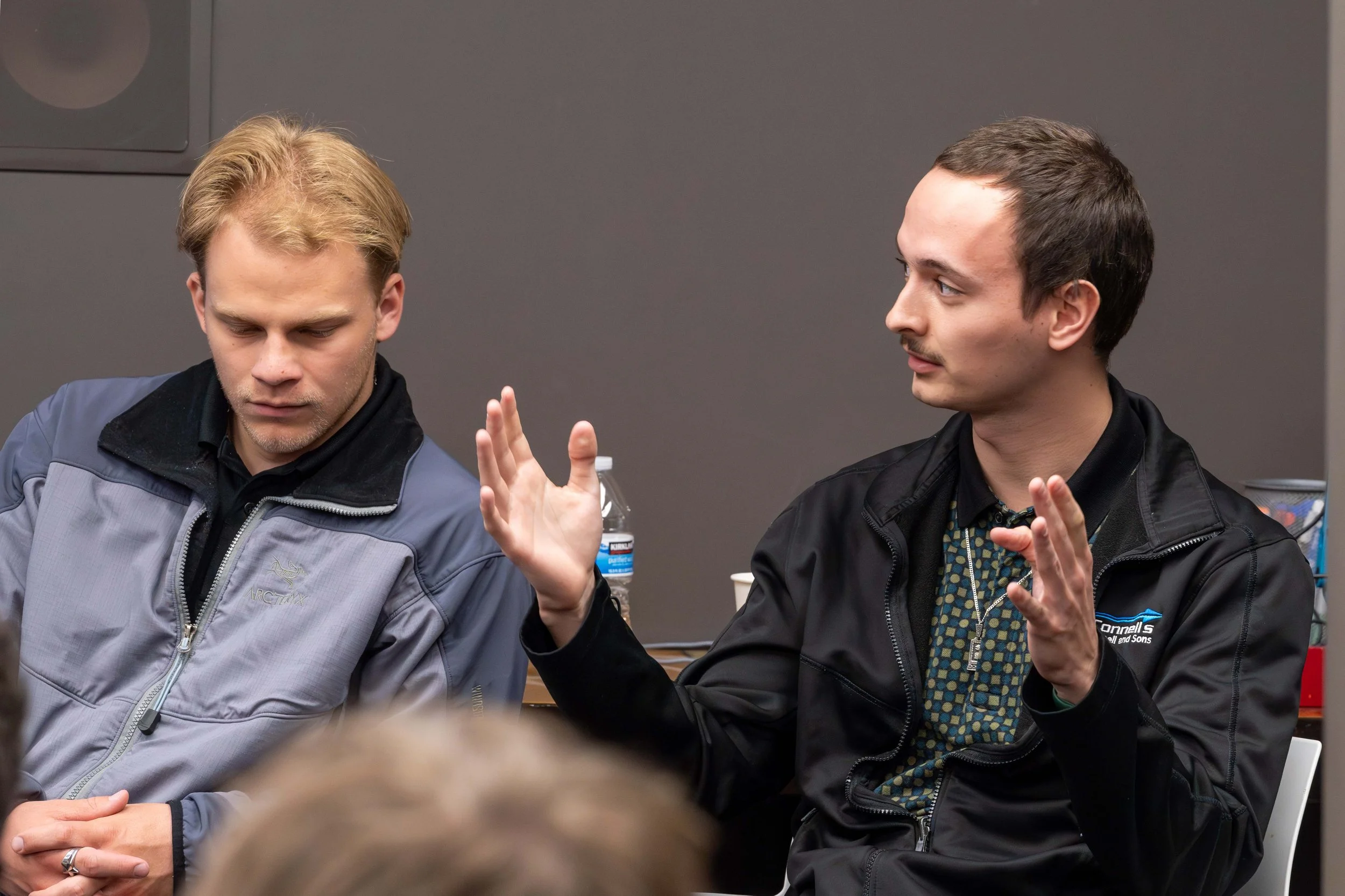 Two men sitting and having a conversation, one with blonde hair and the other with dark hair, in an indoor setting with a gray wall and a water bottle in the background.
