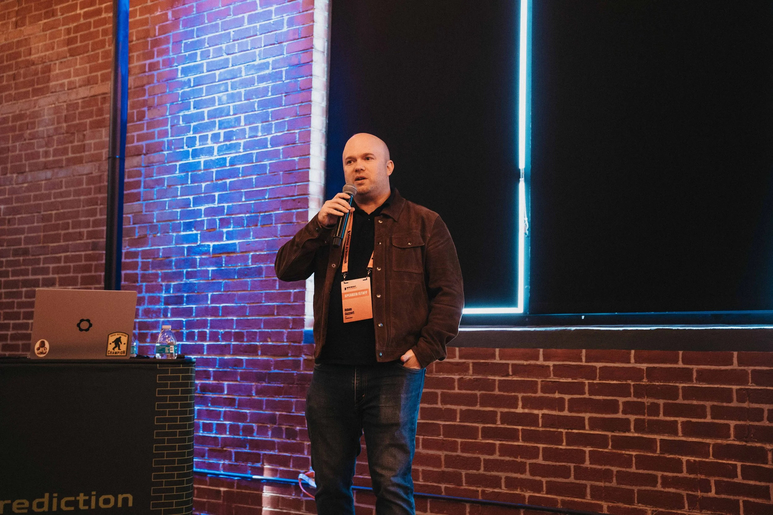 A man speaking into a microphone during a conference or presentation, standing in front of a brick wall with blue lighting accents, with a laptop and water bottle on the table beside him.