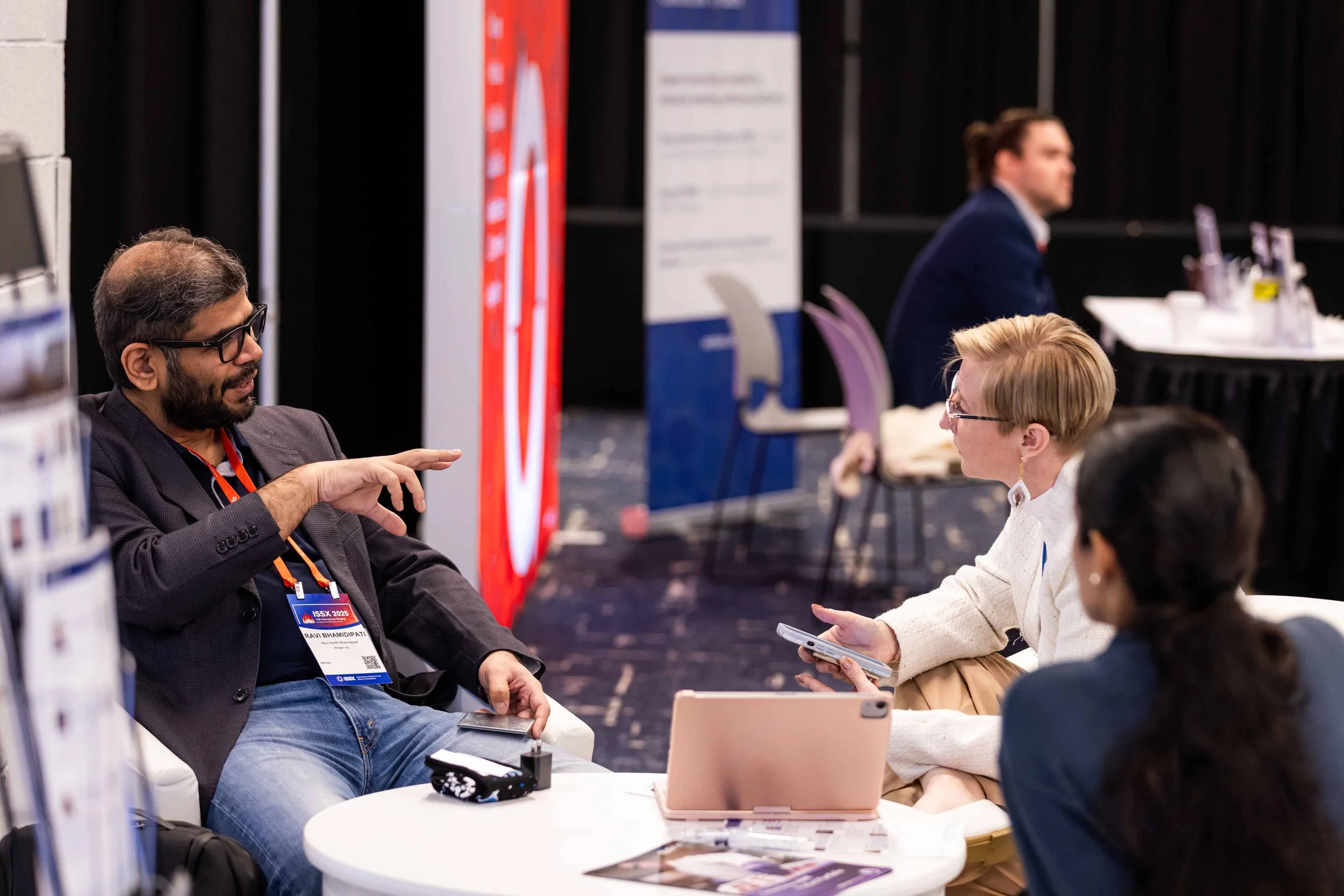 Four people sit at a round white table engaged in conversation at a professional event. The man on the left with glasses and a beard is speaking, while the woman across from him, holding a phone, listens. Two women, one with dark hair and the other w