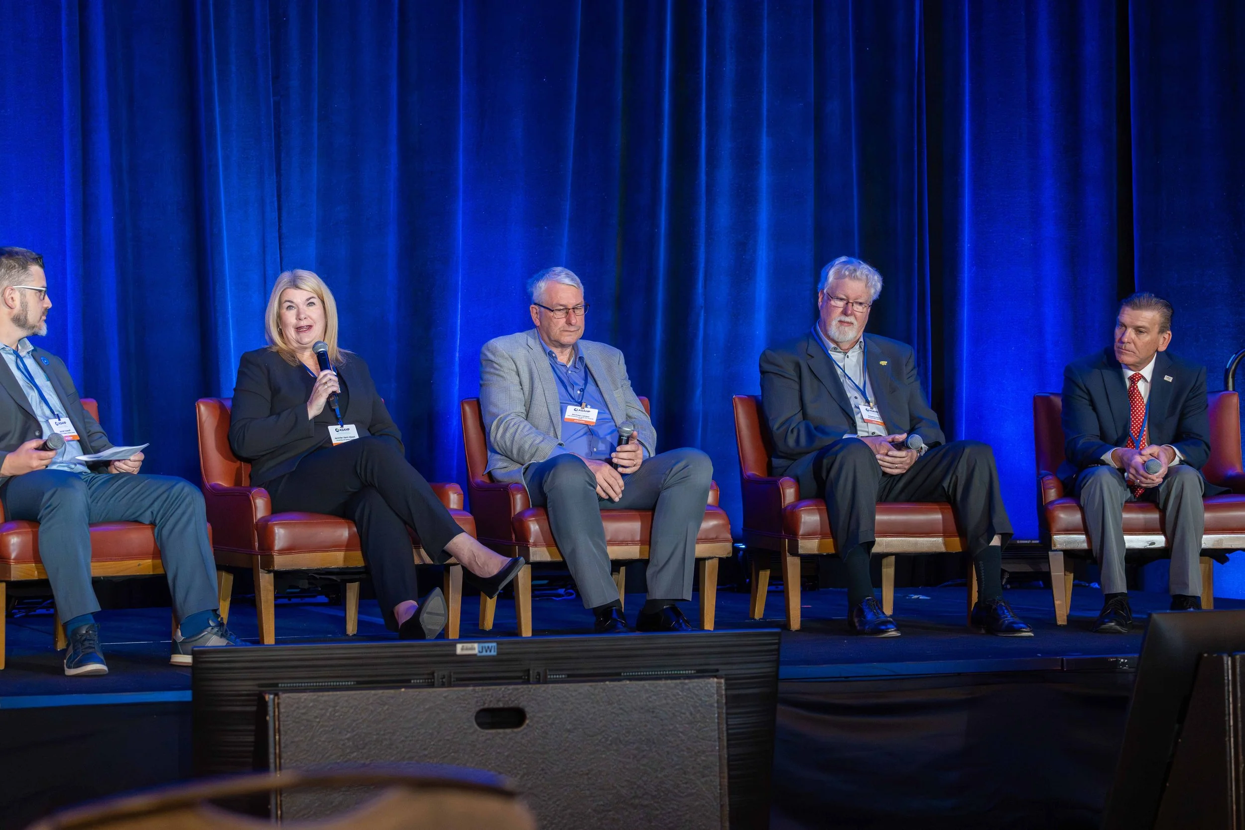 Five people sitting on stage chairs in a panel discussion, with a woman speaking into a microphone, against a blue curtain background.