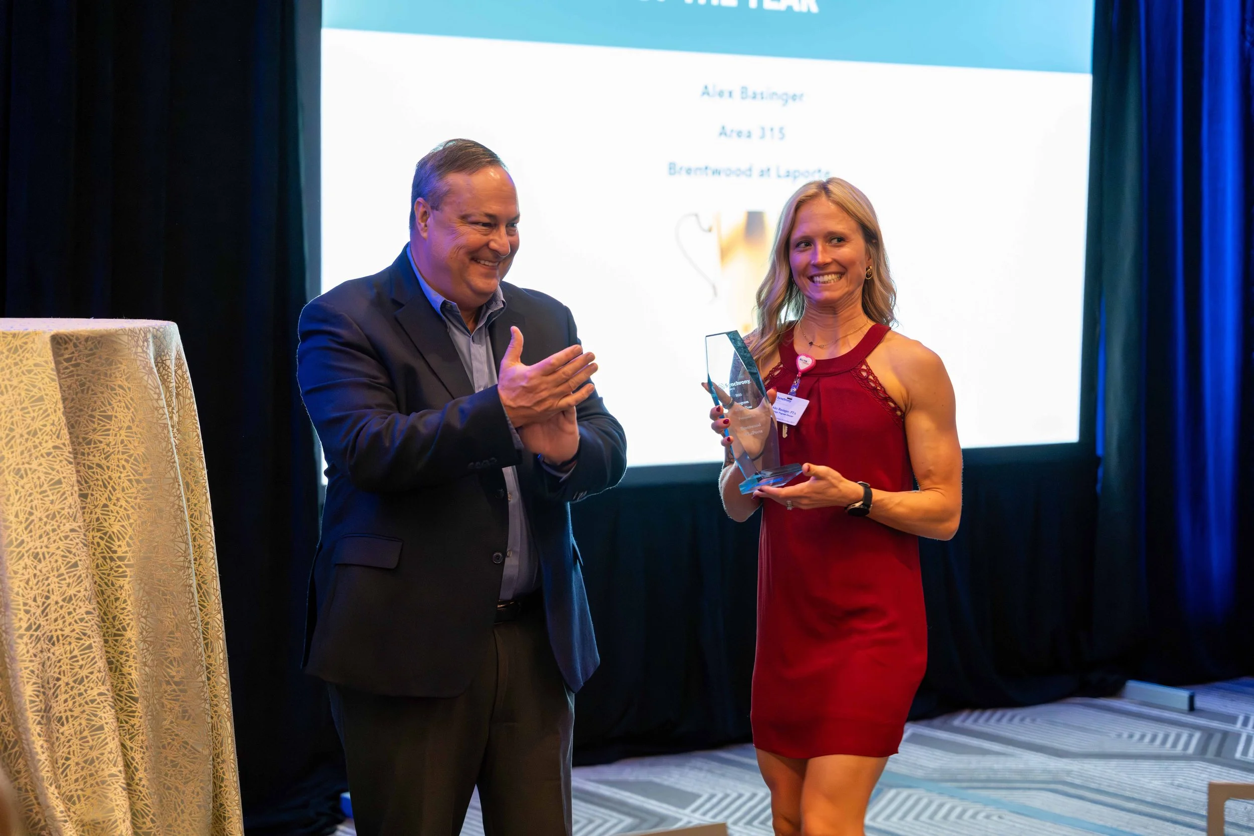 A woman in a red dress receiving an award from a man in a suit on stage with a presentation screen in the background