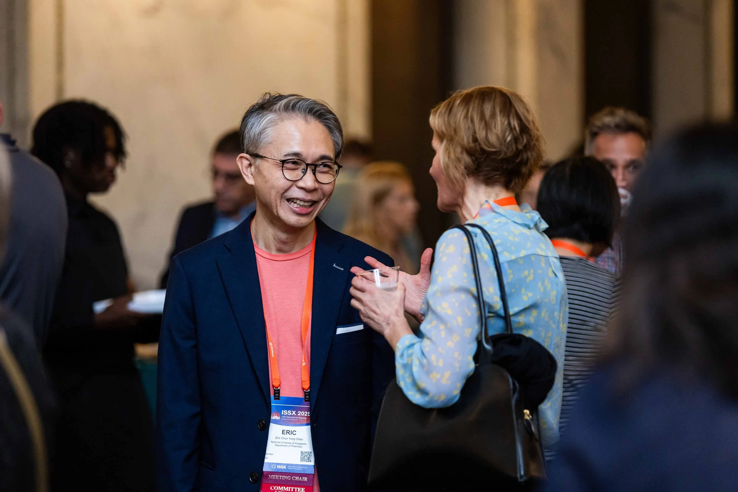 Two people engaged in conversation at a professional conference, with one person wearing a dark suit jacket and glasses, and the other wearing a floral blouse with a black shoulder bag, in a crowded indoor setting.
