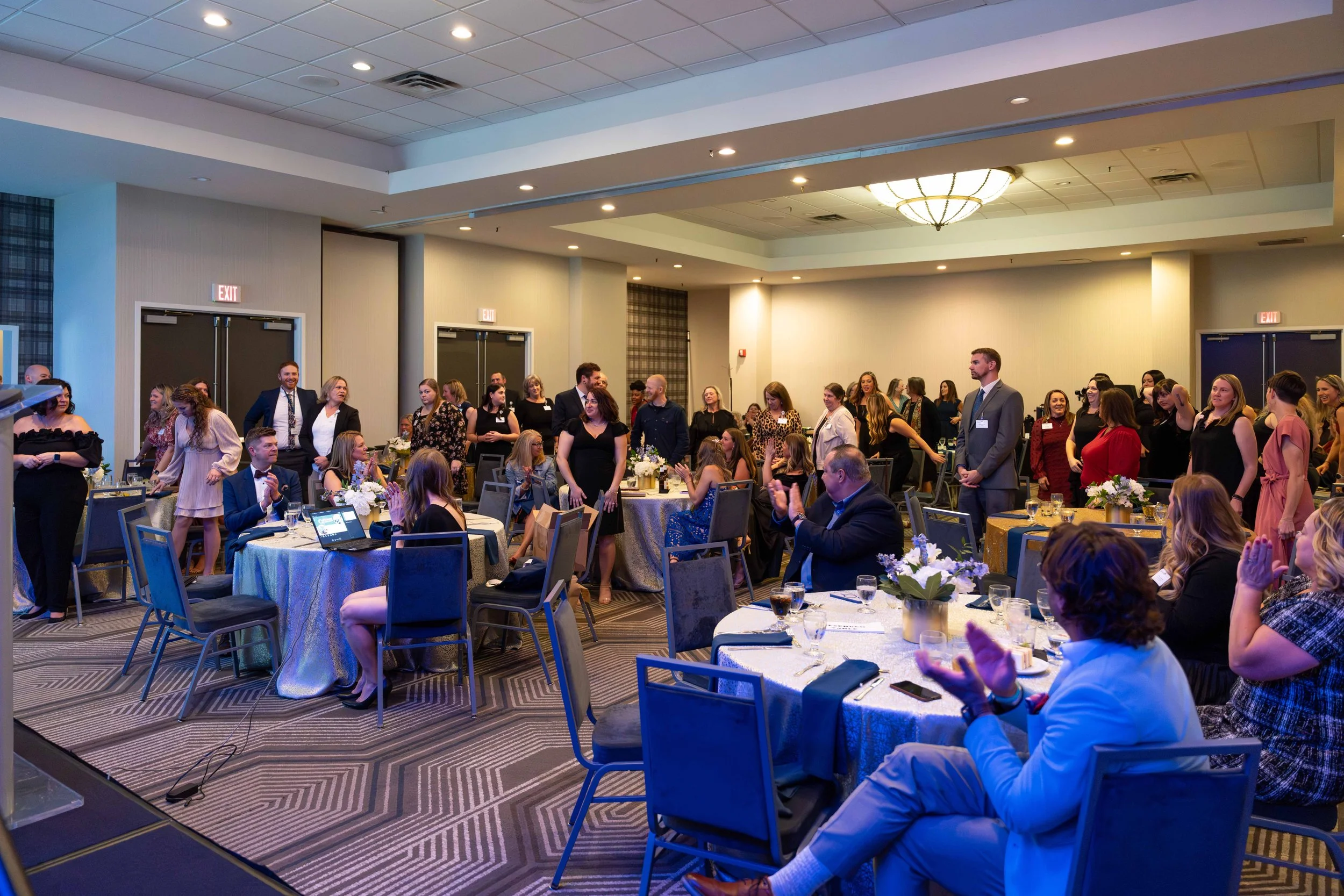 People standing and sitting at tables in a banquet hall, some clapping, others talking, with a formal event taking place.