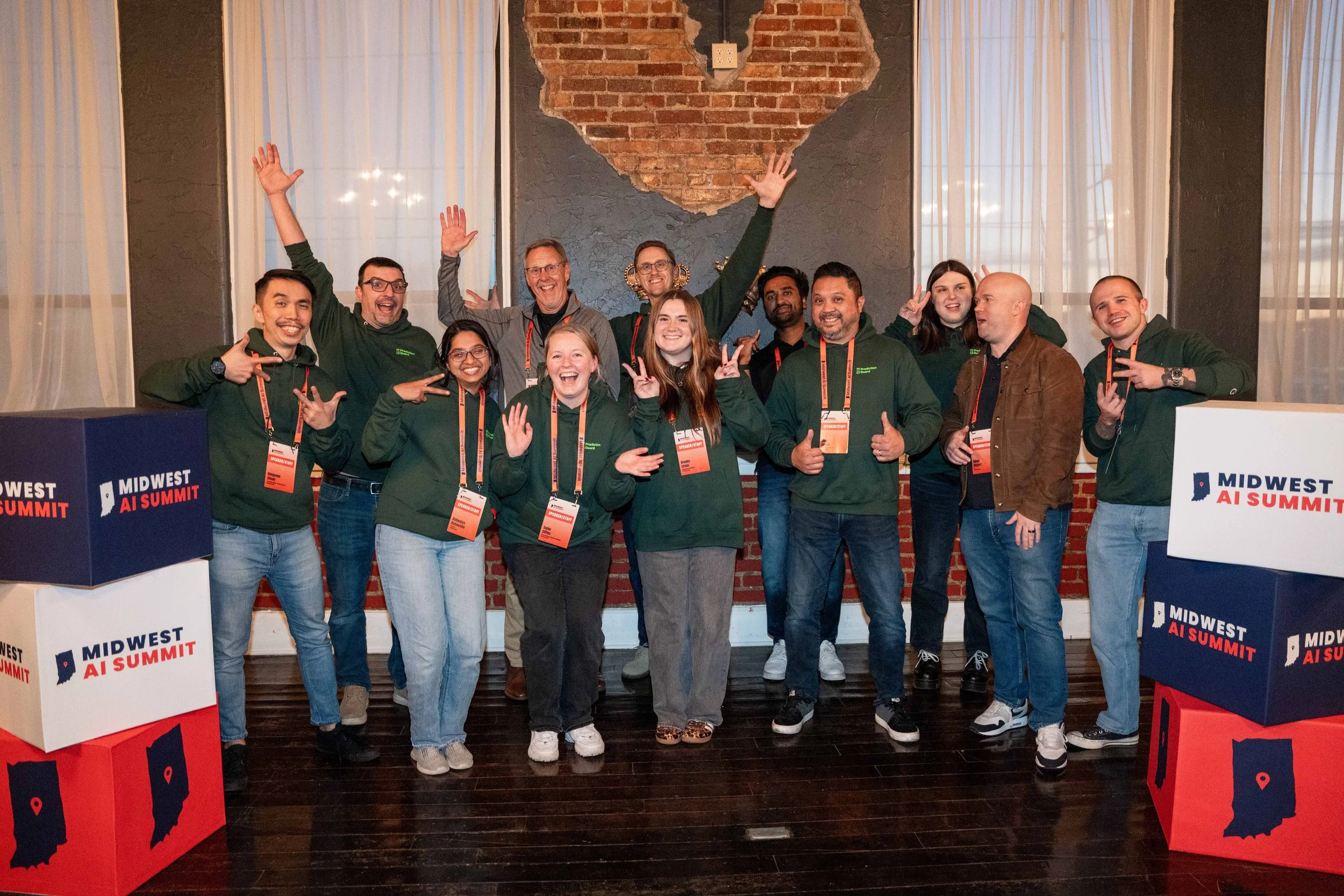 Group of people at the Midwest AI Summit, posing and smiling, with some making peace signs. They are wearing dark green hoodies, lanyards, and standing indoors in front of a brick wall with a heart-shaped opening and logos of the event.