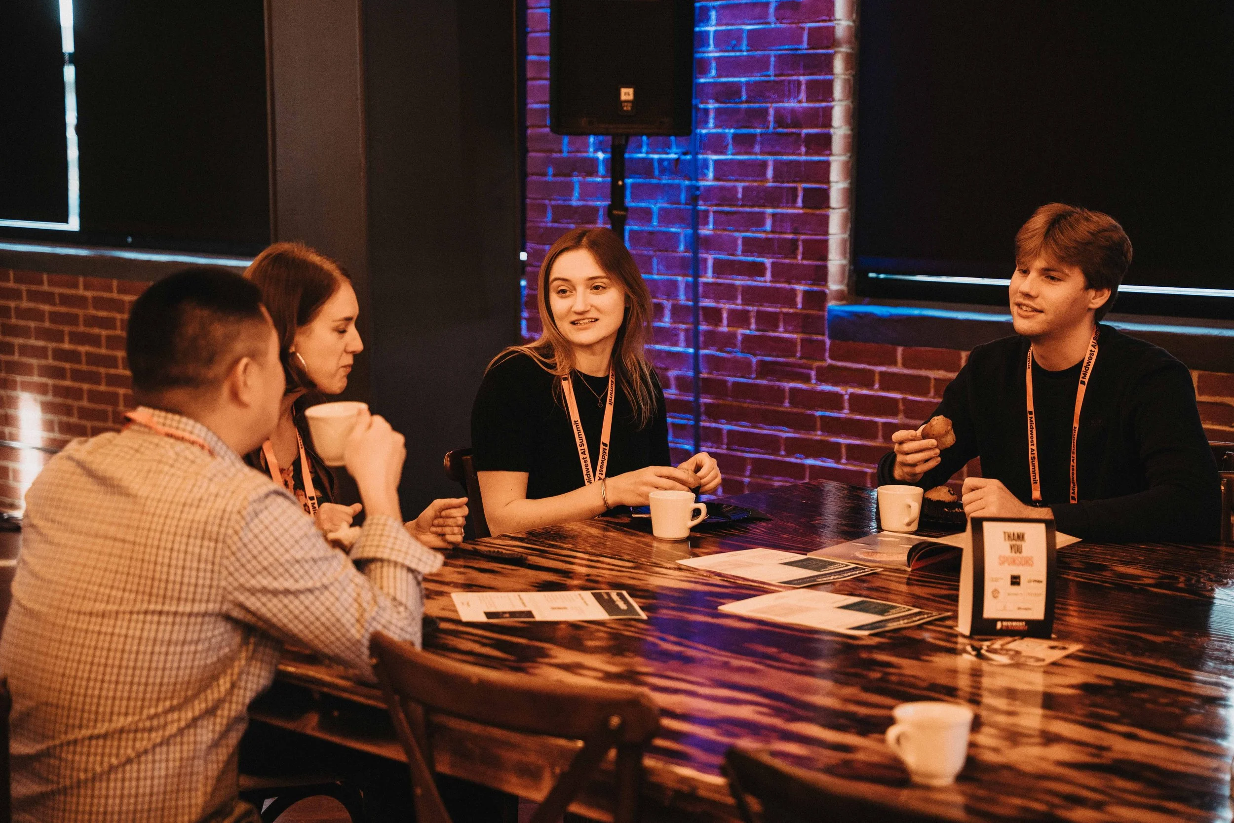Group of four young adults sitting at a long wooden table in a dimly lit room with exposed brick walls, engaging in conversation with coffee cups and papers on the table.