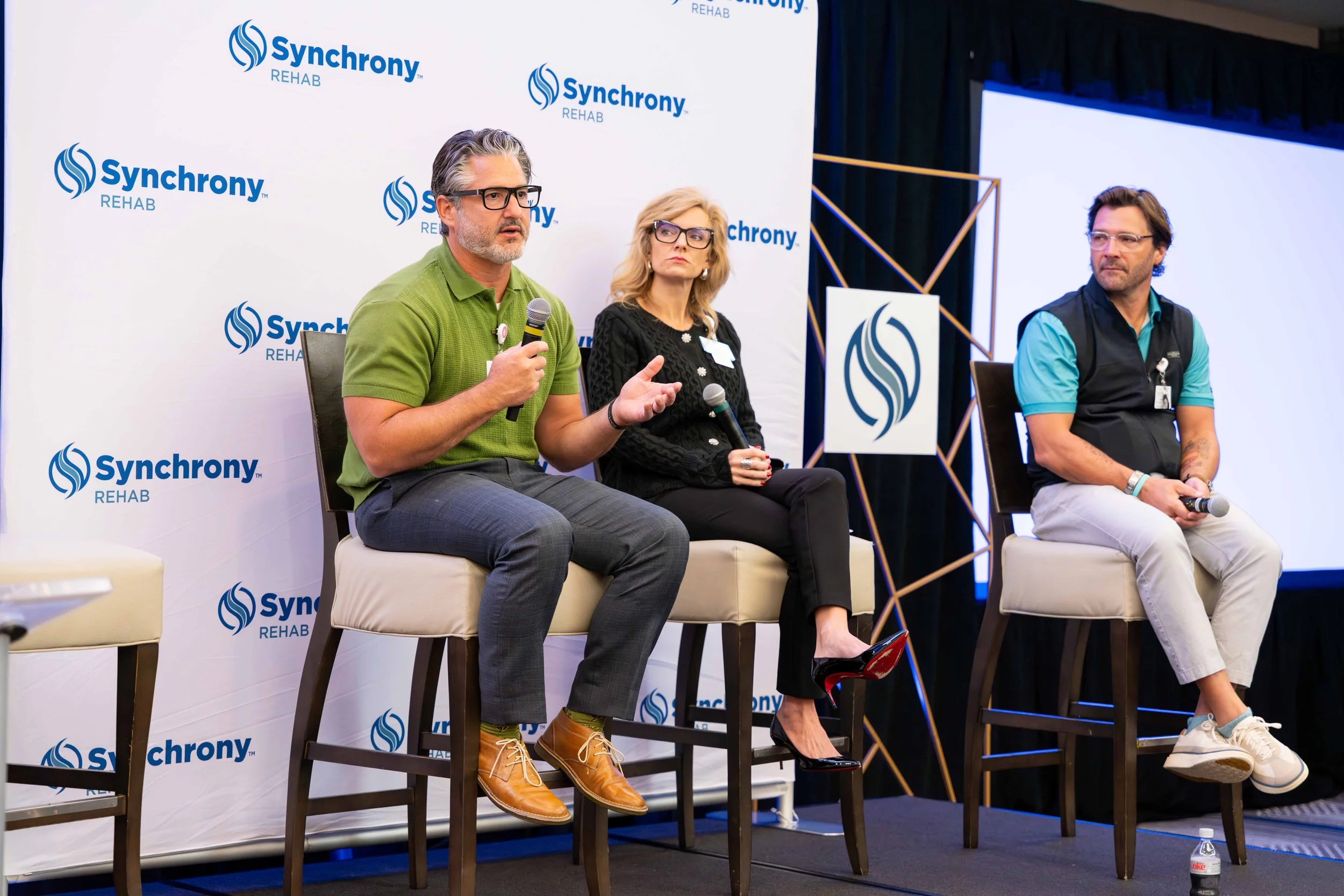 Three people sitting on stage at a Synchorny Rehab event, engaging in a panel discussion. The man on the left is speaking into a microphone, the woman in the middle and the man on the right are holding microphones. The background displays the Synchor