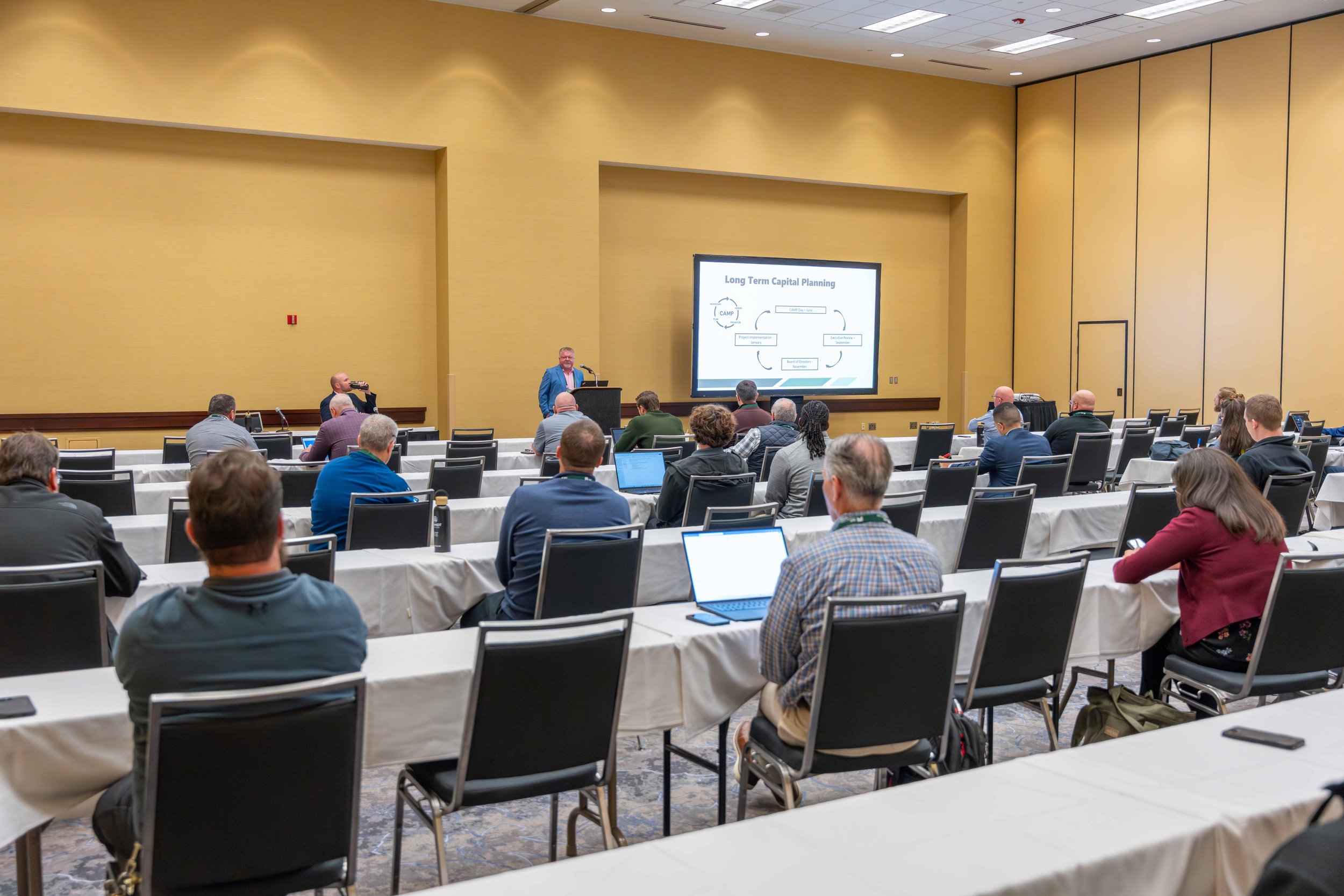 A conference room with attendees sitting at tables, listening to a speaker presenting a slide titled 'Long Term Capital Planning'.