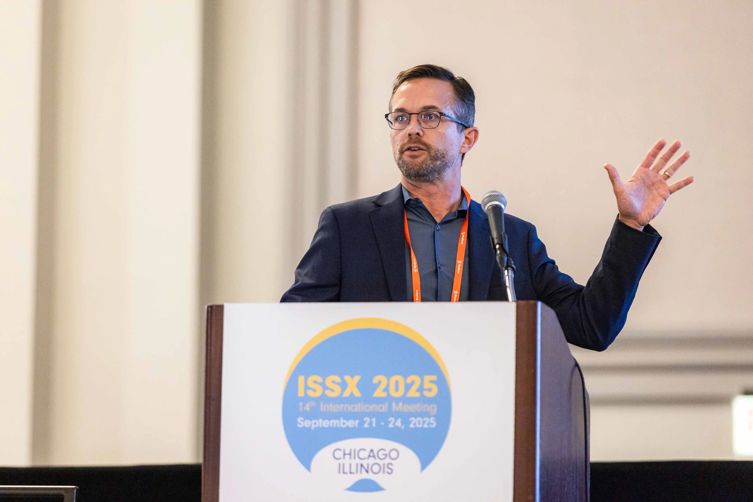 A man with glasses and a beard wearing a dark suit and blue shirt, standing at a podium, gesturing with his right hand while speaking into a microphone at the ISSX 2025 conference in Chicago, Illinois.