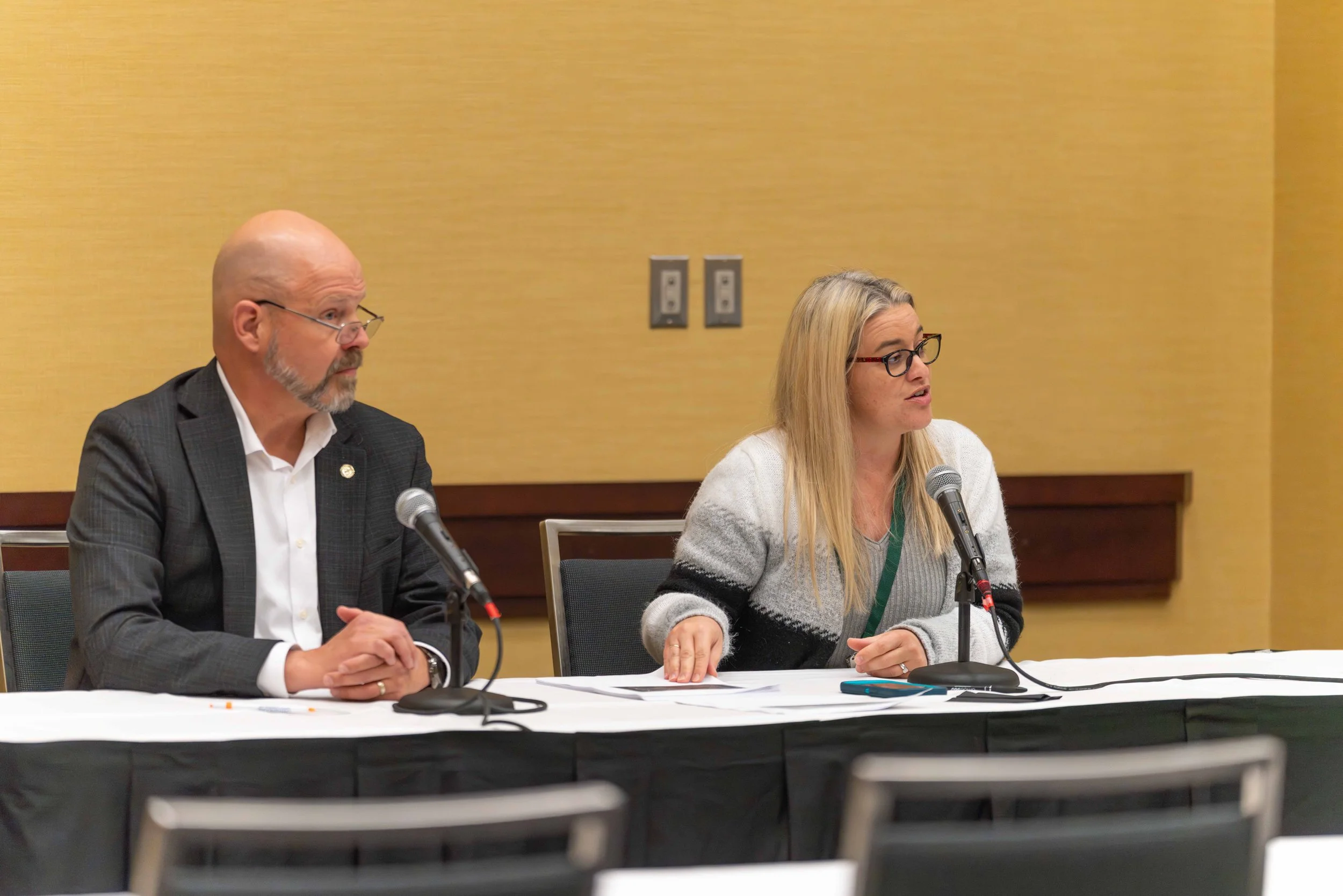 Two individuals sitting at a table with microphones during a conference or panel discussion, with a yellow wall in the background.