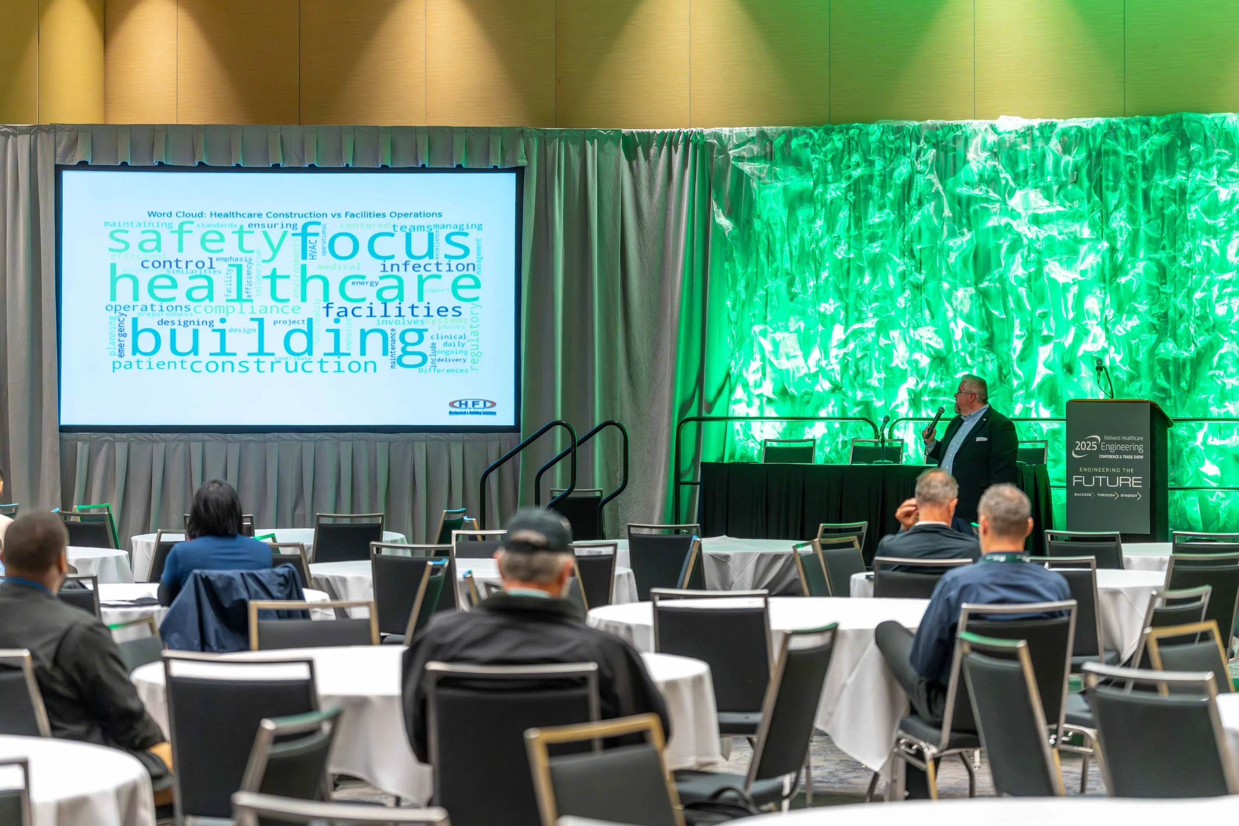 A conference room with round tables and chairs, a speaker standing at a podium, and a large screen displaying a word cloud about healthcare and building.