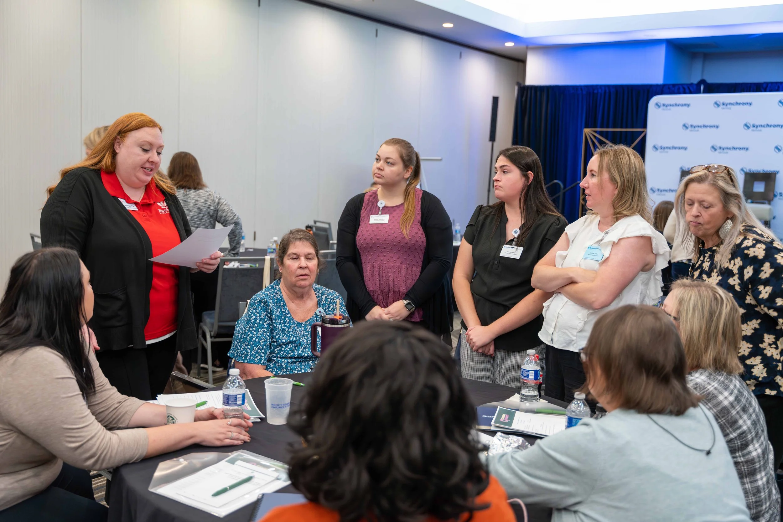 A woman in a red shirt and black jacket is speaking to a group of women seated around a table at a conference. The women are listening attentively, some with arms crossed or hands folded, and the table has water bottles, notebooks, and documents. The