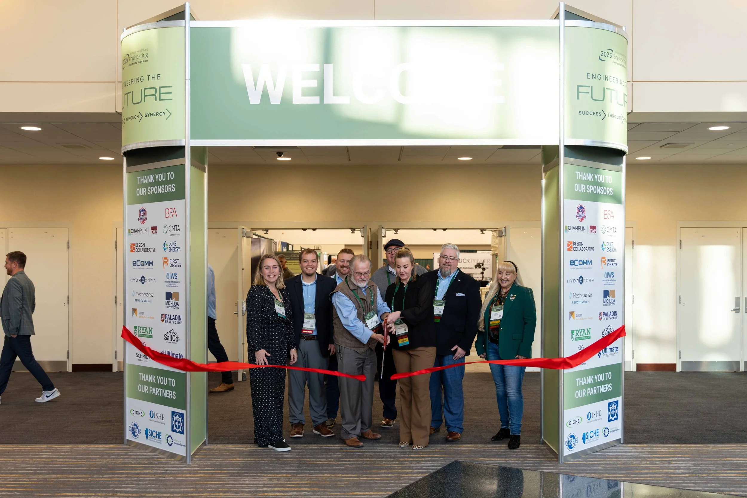 Group of people at a ribbon-cutting ceremony at an event entrance, with a large green welcome sign overhead and sponsor logos on the side panels.
