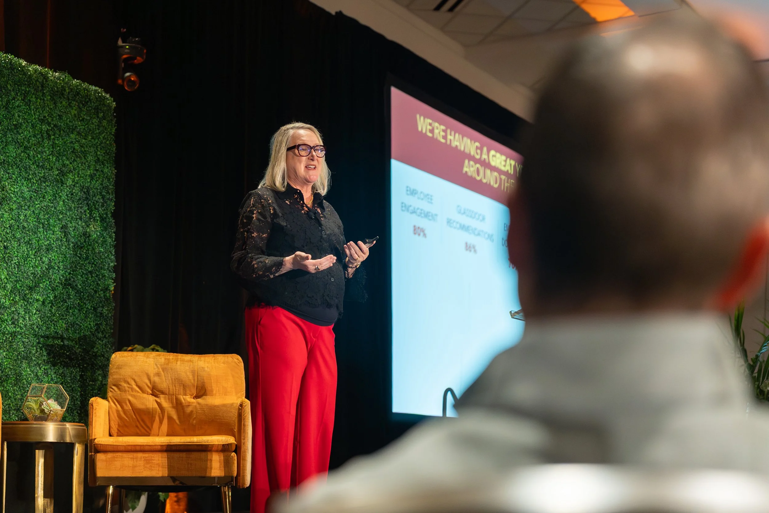 A woman in black lace blouse and red pants giving a presentation on stage, with a large screen displaying a slide behind her, and audience members in the foreground.