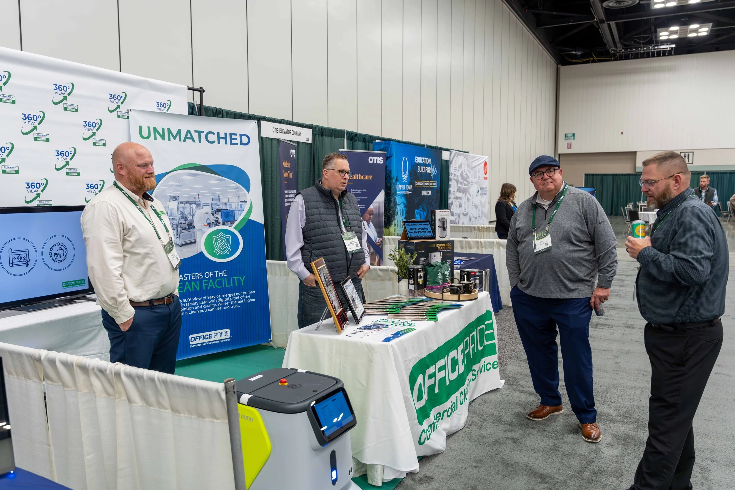 Four men are standing and talking at an exhibition booth. The booth displays promotional materials for Office Pride Commercial Cleaning Services, with banners and a table. One of the men is holding a cup and smiling, while the others are engaged in c