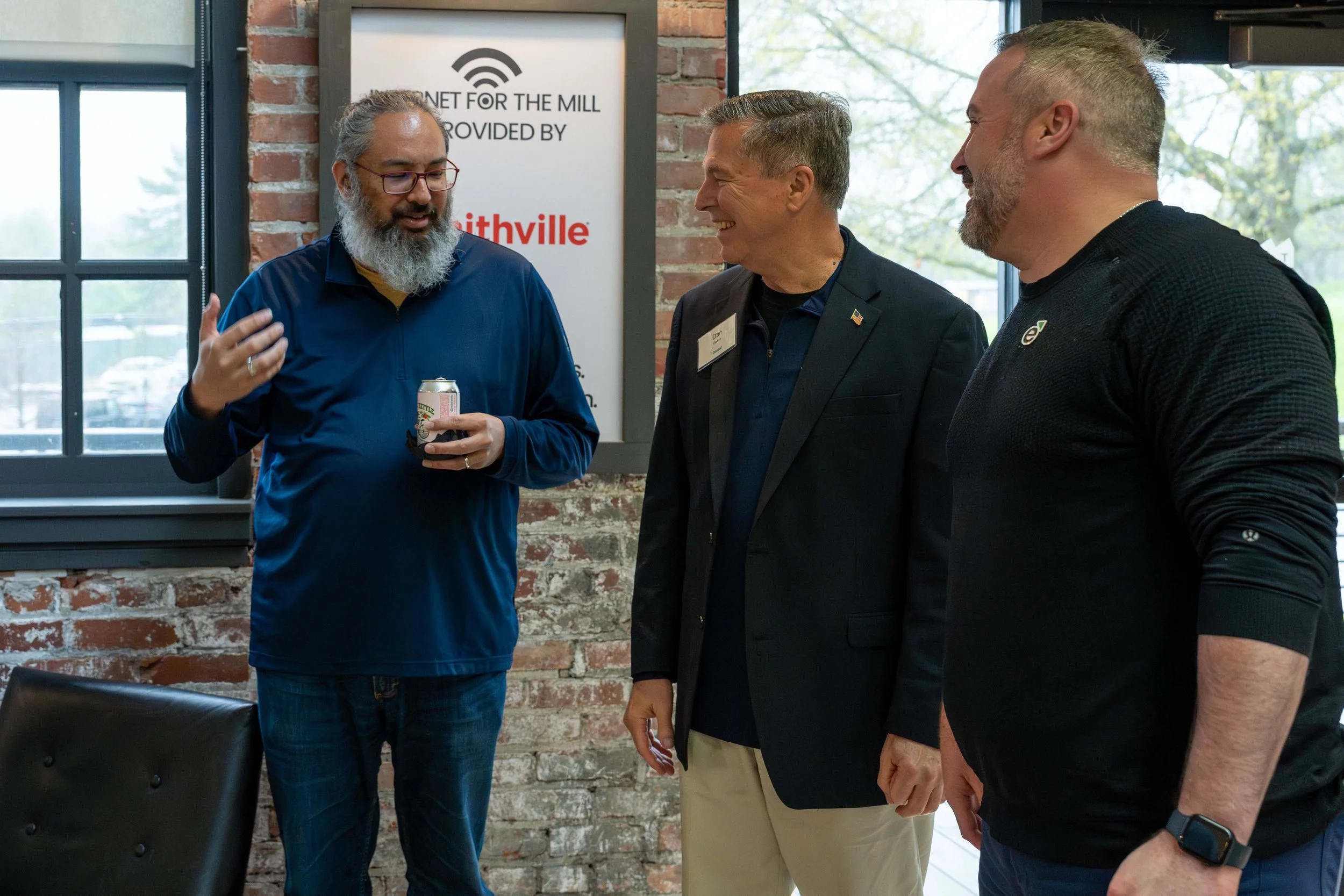 Three men having a conversation indoors near a brick wall and large windows, with a sign behind them that says 'Internet for the Mill provided by Smithville'.