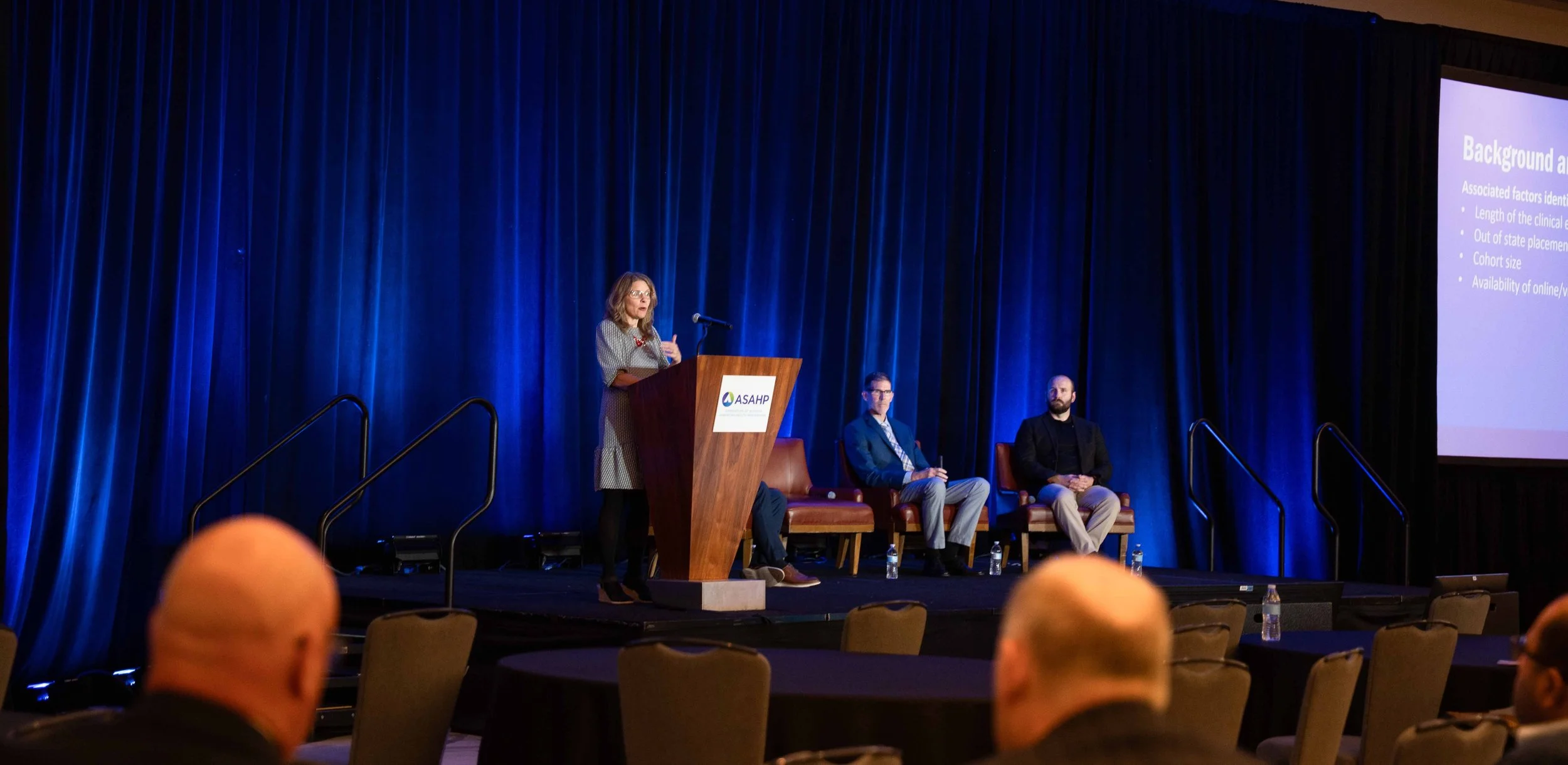 A woman standing at a podium giving a presentation at an indoor conference event. There are two men sitting on stage behind her, one with glasses and a dark blazer, and another with a beard and black blazer. The backdrop is a blue curtain, and a larg