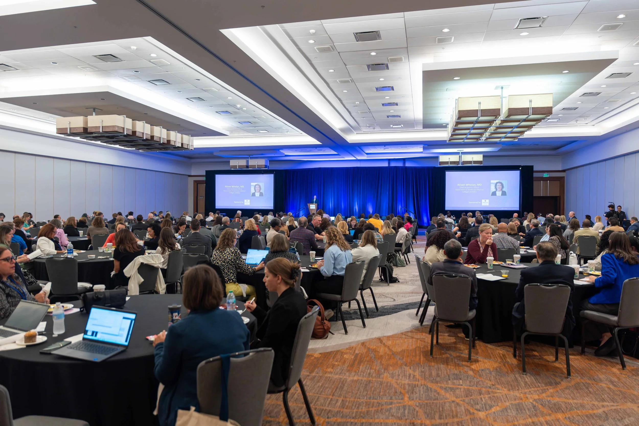Large conference room filled with attendees sitting at round tables, listening to a speaker on stage with two large screens displaying a presentation.