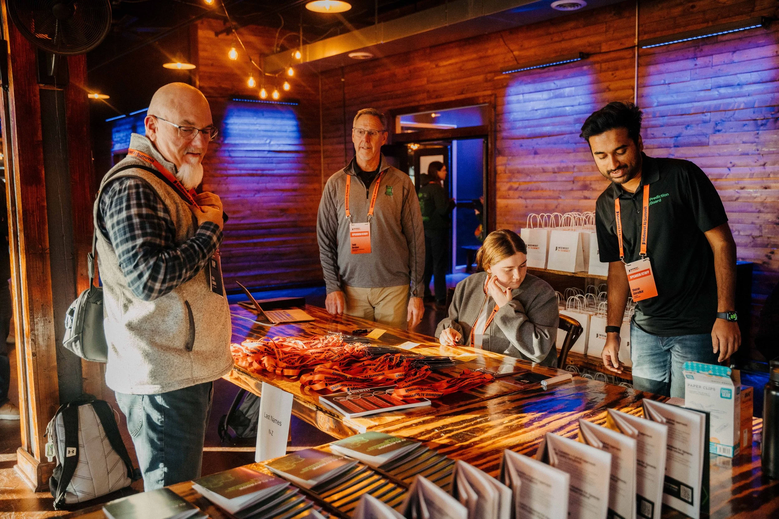 People attending an event at a registration desk with name tags, brochures, and gift bags in a wooden-finished room with warm lighting.