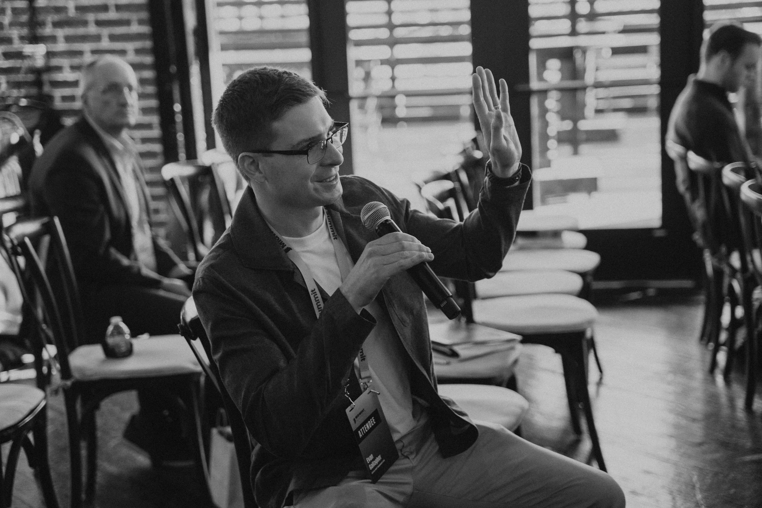 A young man with glasses and a short haircut, sitting in a chair, holding a microphone in his right hand and raising his left hand, possibly asking a question or making a comment at a conference or seminar. He is wearing a lanyard with an attendee ba