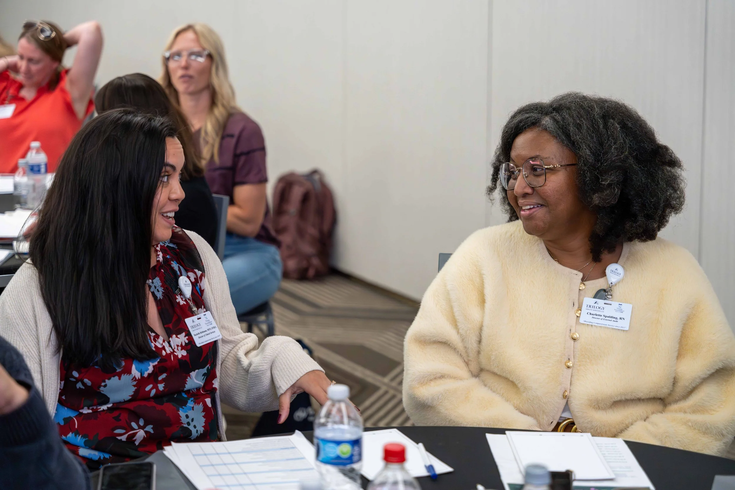 Two women seated at a table engaged in conversation, with several other women in the background, at a professional or conference setting.