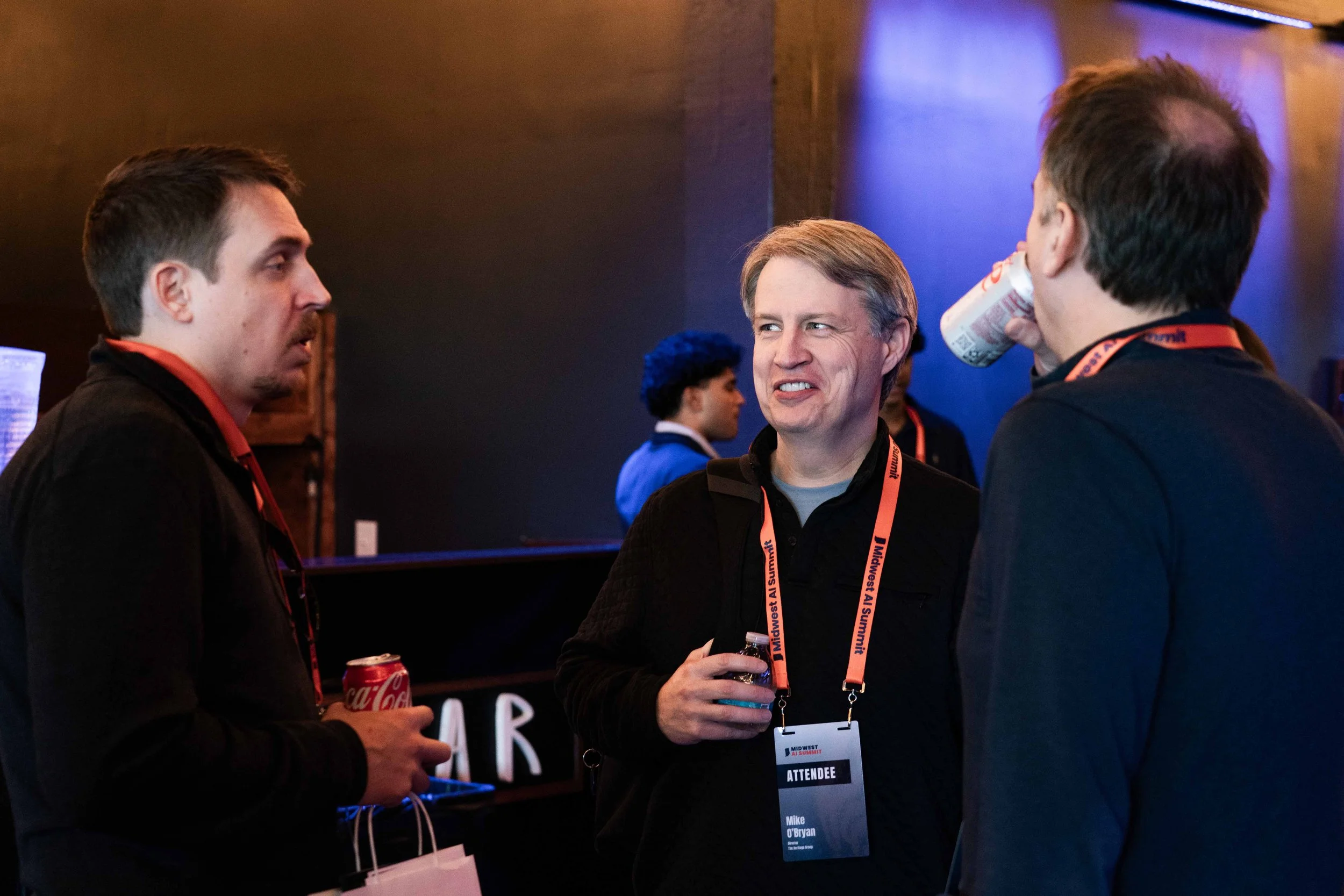 Three men are engaged in conversation at a conference, wearing conference badges and lanyards. One man is holding a soda can and a bag, another is holding a drink, and they are in a dimly lit room with a bar in the background.