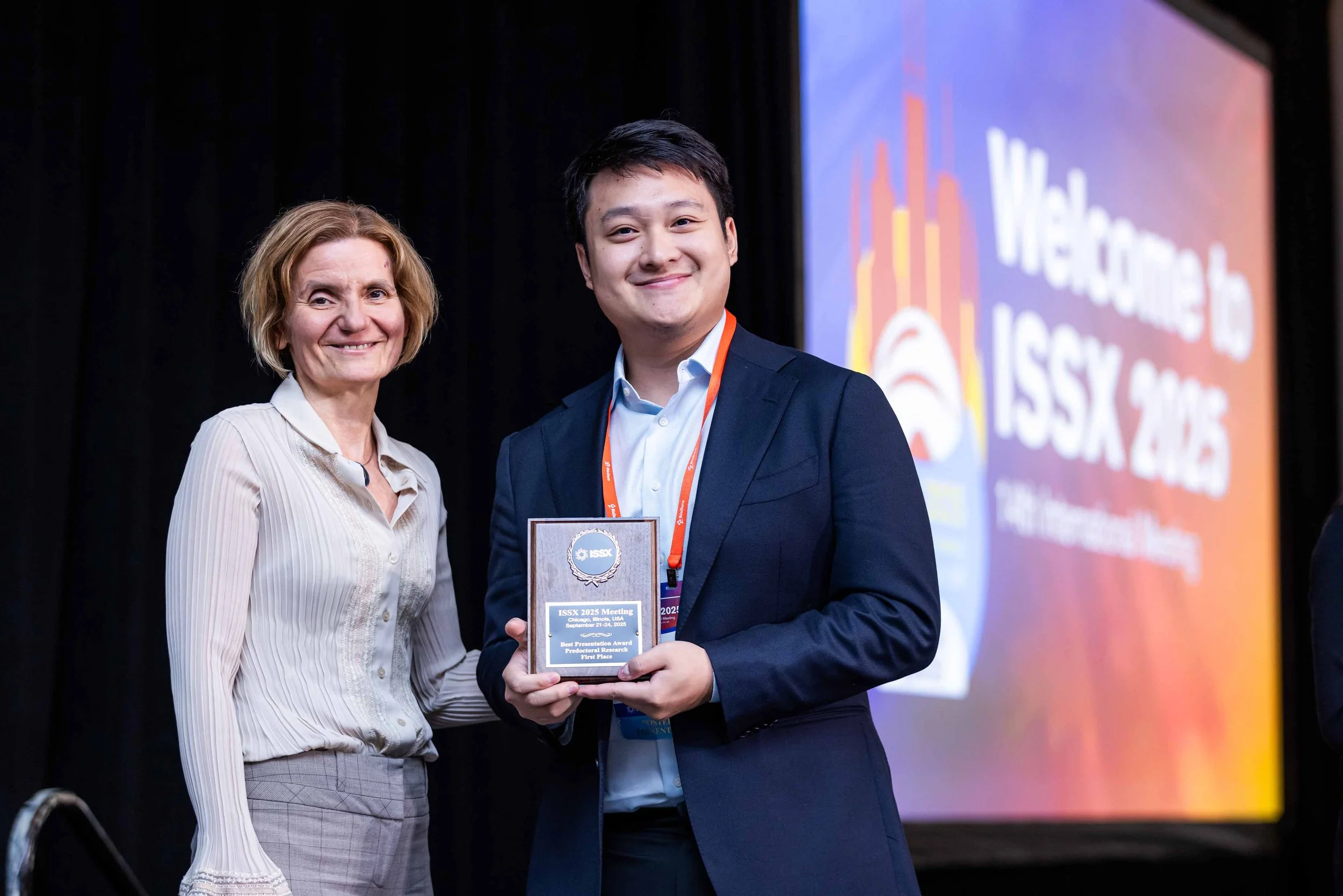 A young man in a navy suit holds a plaque and smiles, standing next to an older woman in a light-colored blouse and plaid skirt, at a formal event with a large screen displaying 'ISSX 2023' in the background.