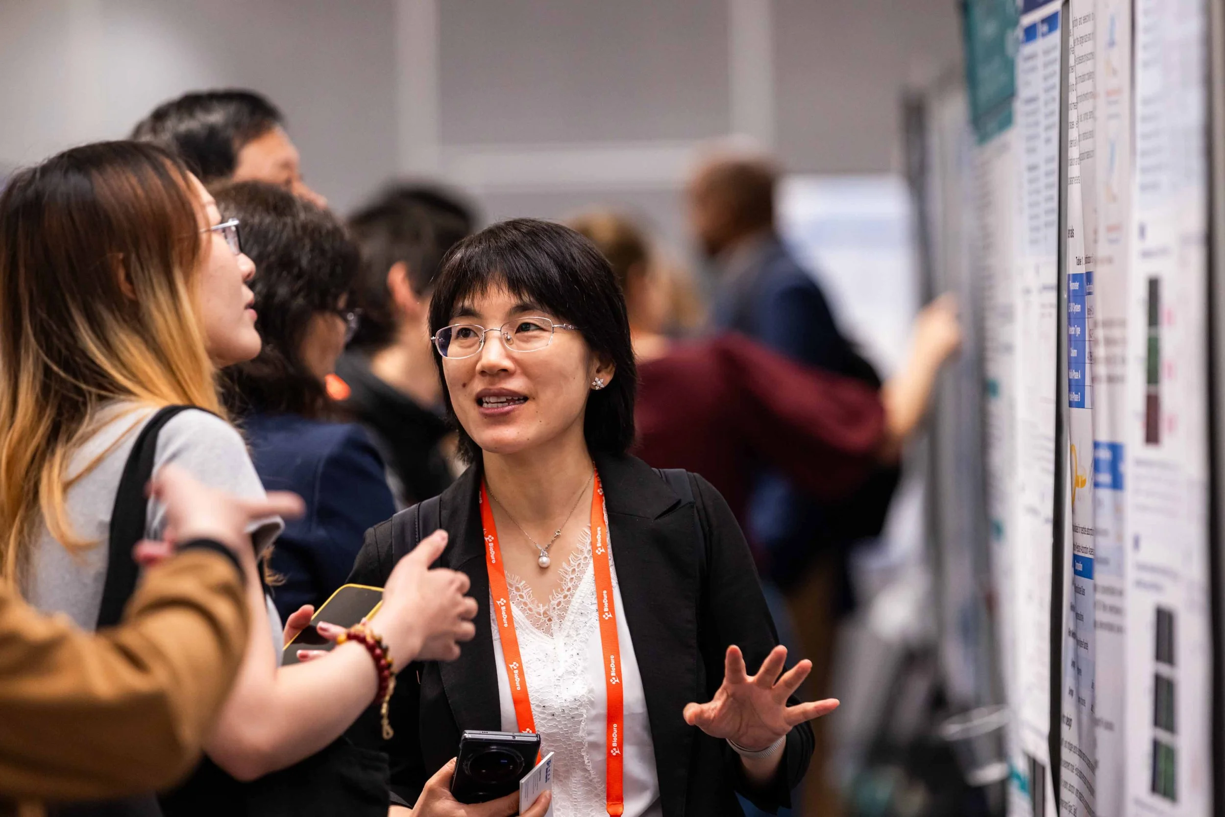 A woman with glasses and short black hair speaking to a group of people at a conference, with research posters on display in the background.