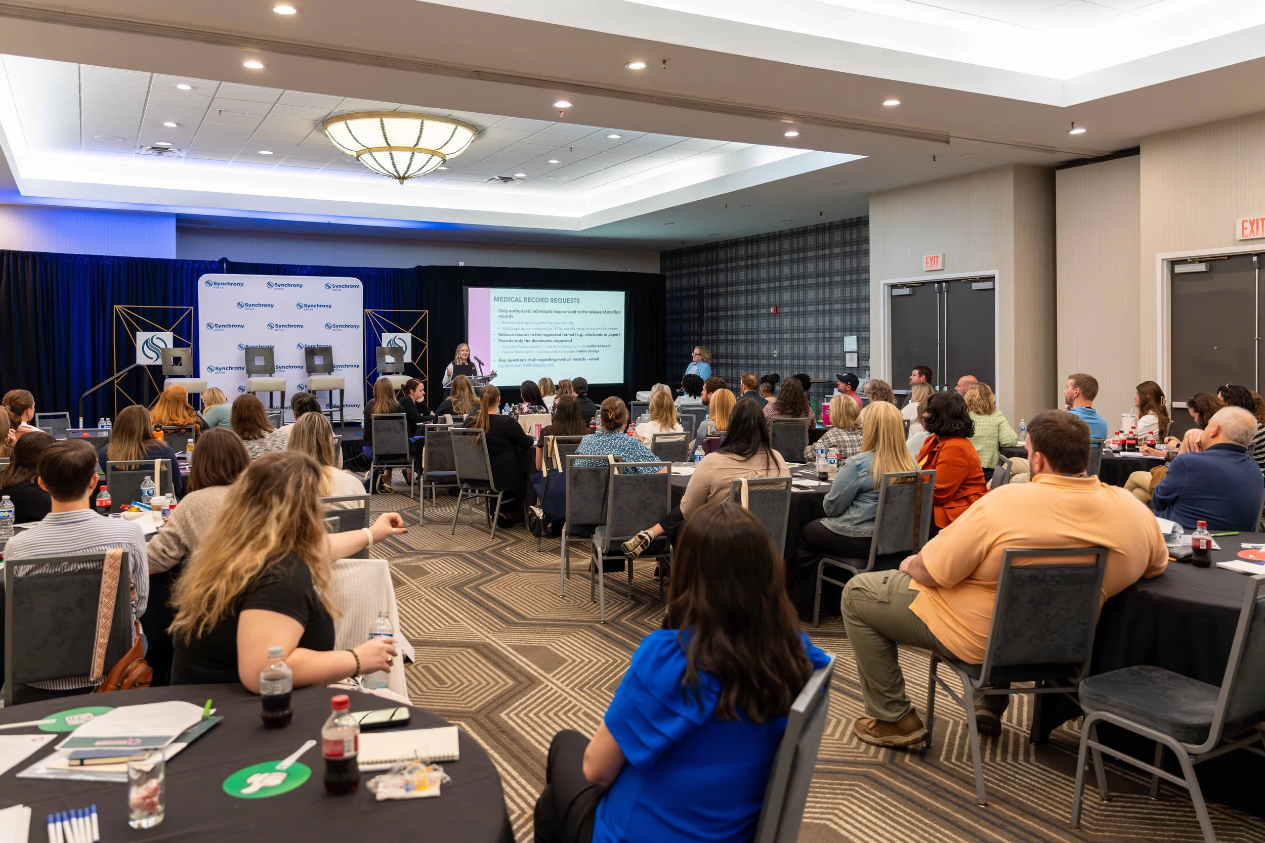A conference room filled with people sitting at round tables, attentively listening to a speaker presenting slides on a large screen. The room has a professional setting with a stage, backdrop, and multiple attendees with notebooks and water bottles 