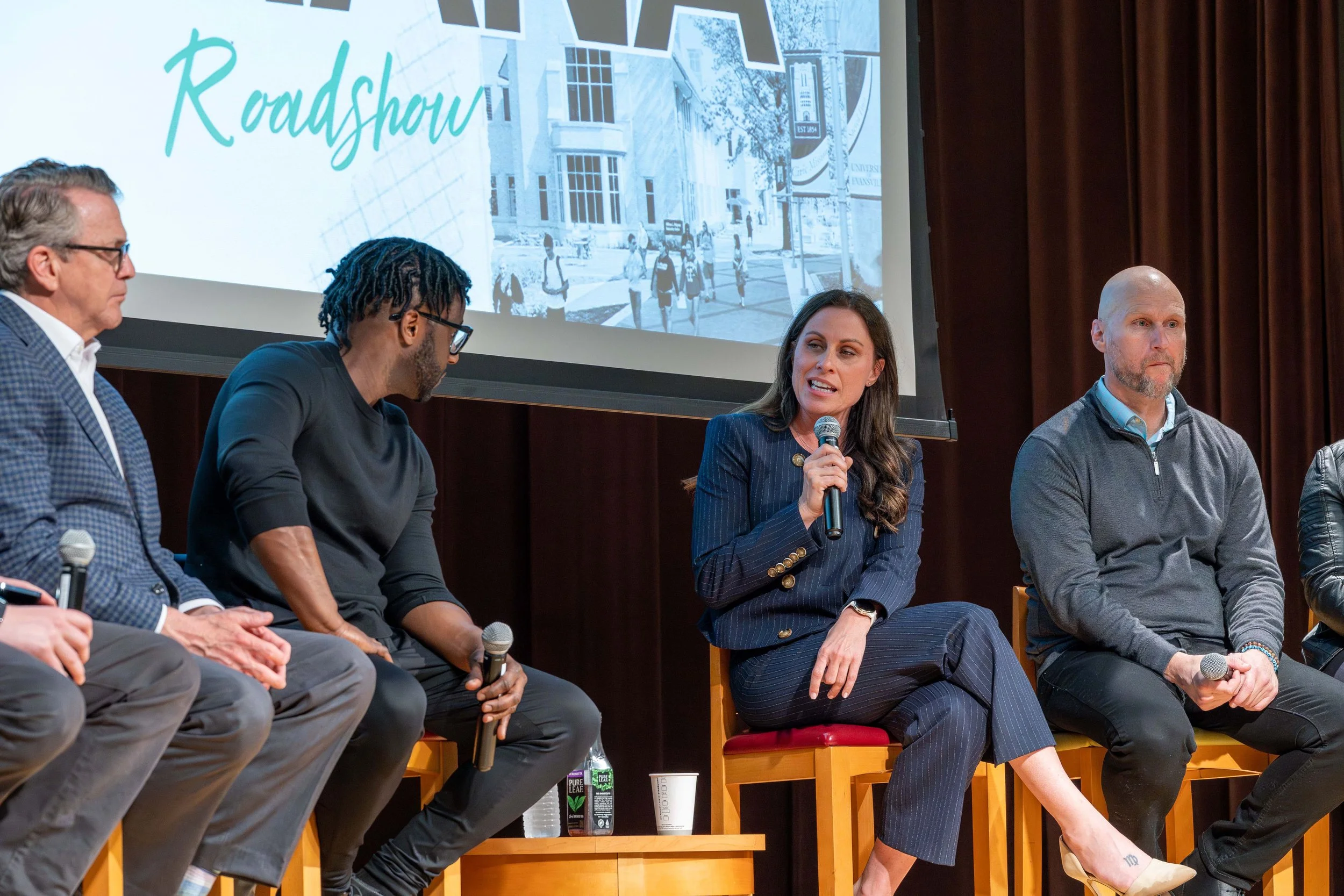 Panel discussion with five people seated on stage, one woman speaking into a microphone, a large screen behind them displaying the word 'Roadshow' and an outdoor campus scene.
