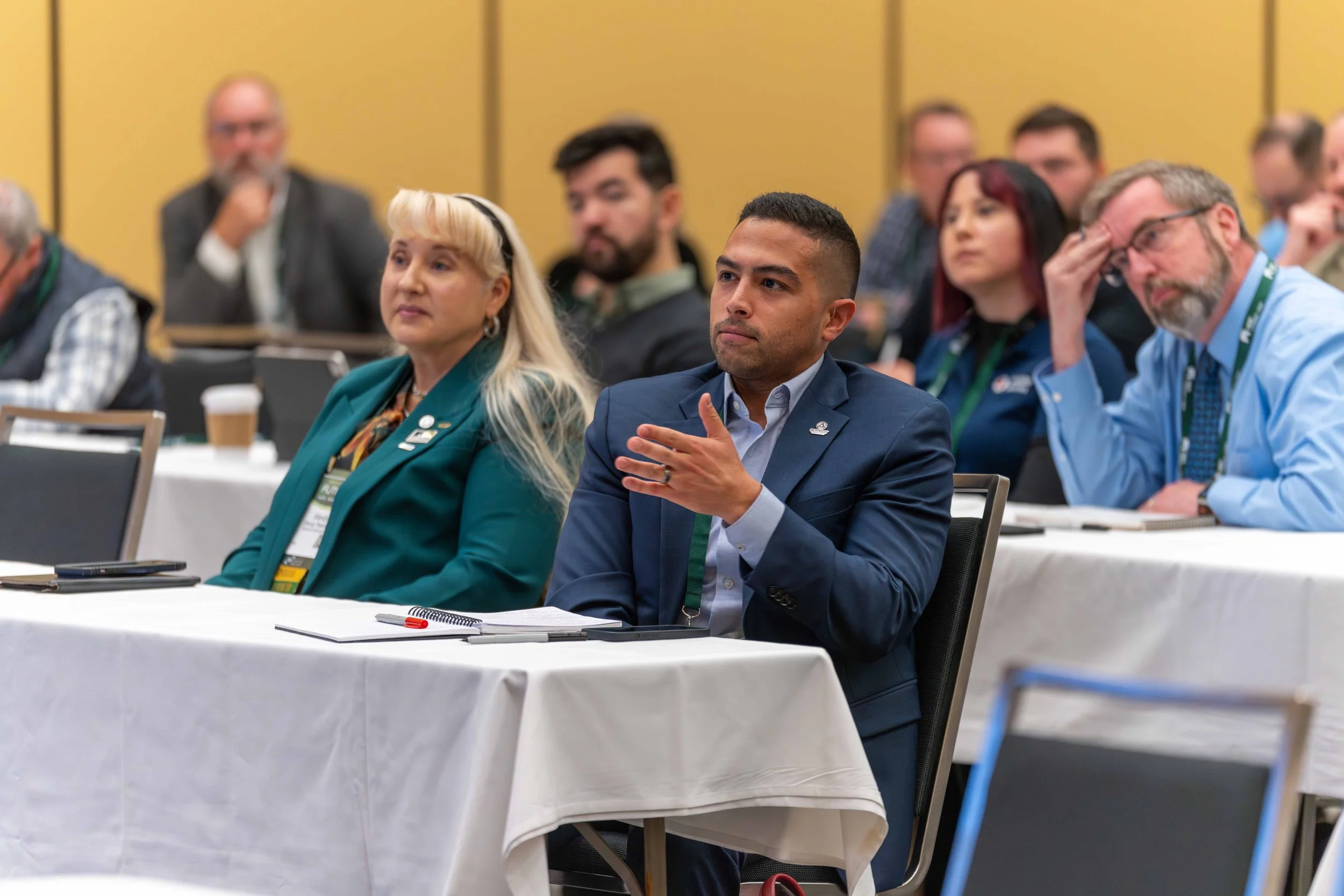 Attentive conference attendees, a man in a blue suit raising his hand, and other adults sitting at long tables with notes and cups in a conference room.