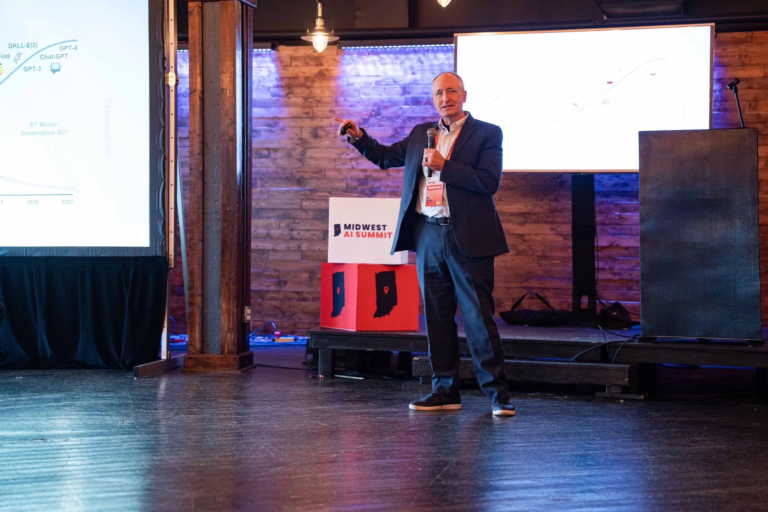 A man in a suit giving a presentation at the Midwest AI Summit, standing on a stage with a microphone in his hand, pointing at a screen showing a graph.
