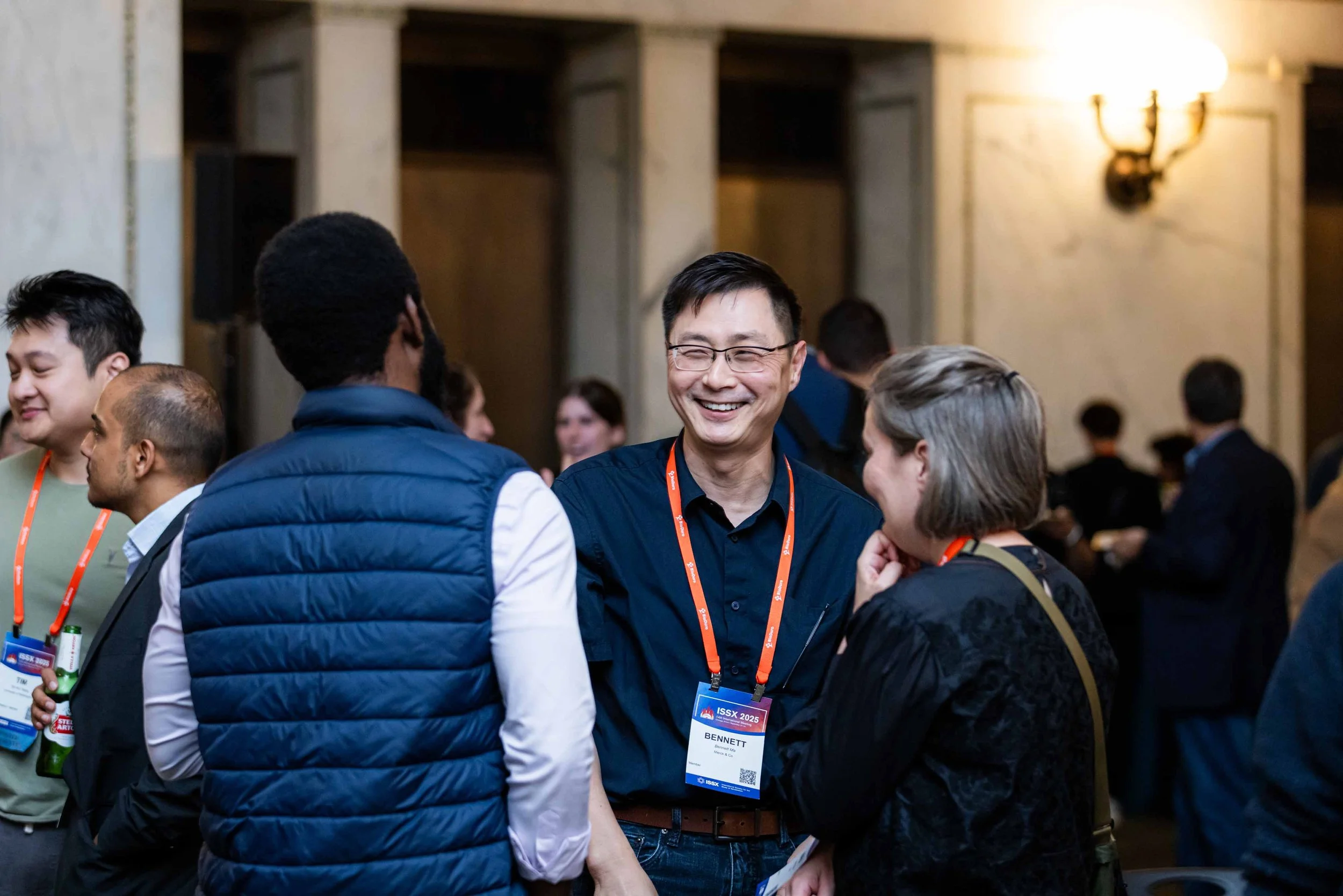People talking and smiling at a conference, wearing name tags, in a well-lit room with ornate walls.