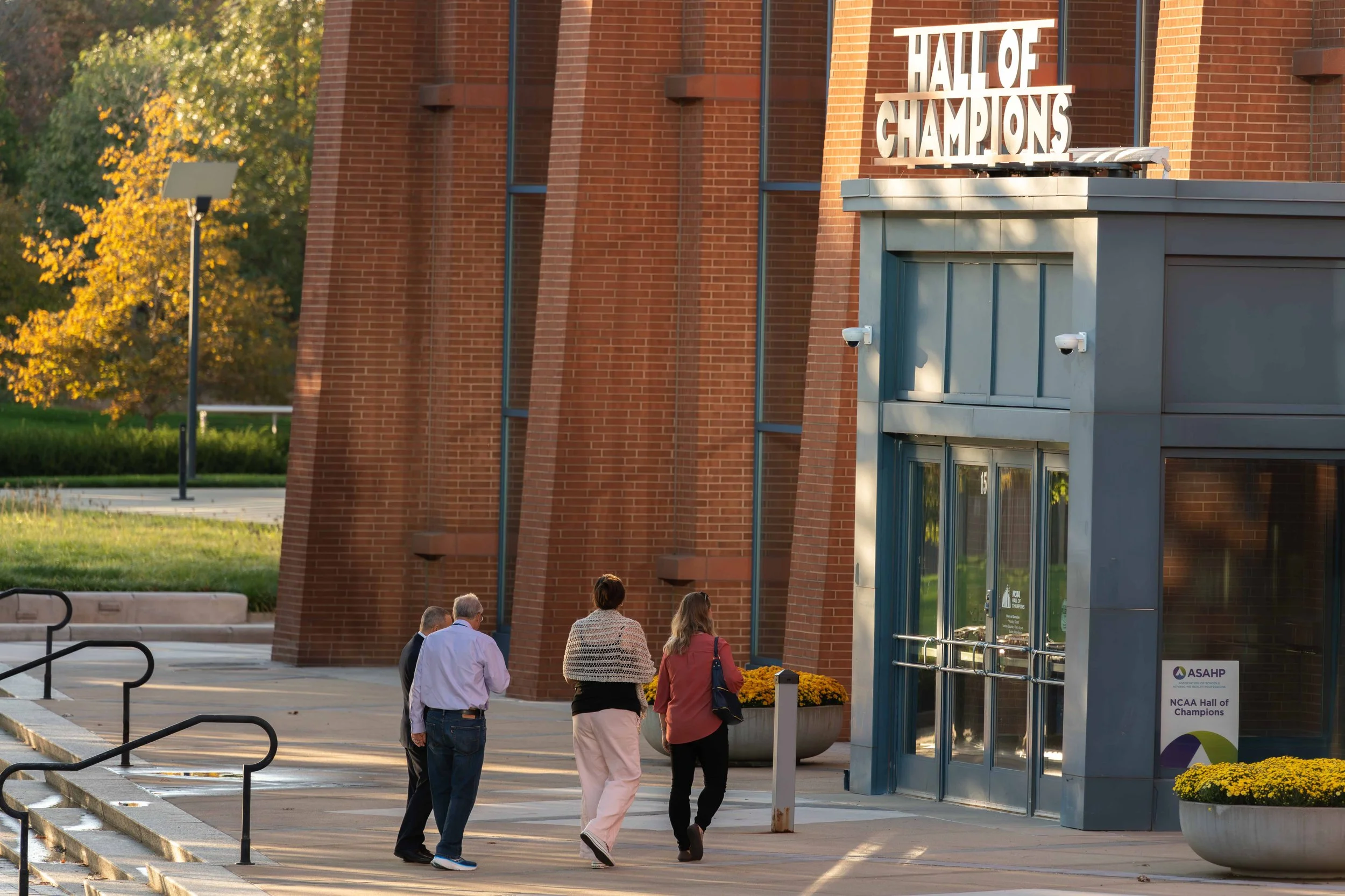 Group of five people walking towards the entrance of a building with a sign that reads 'Halle of Champions.' The building has red brick walls and large glass doors. There are potted yellow flowers outside and a sidewalk with railings. Sunset lighting