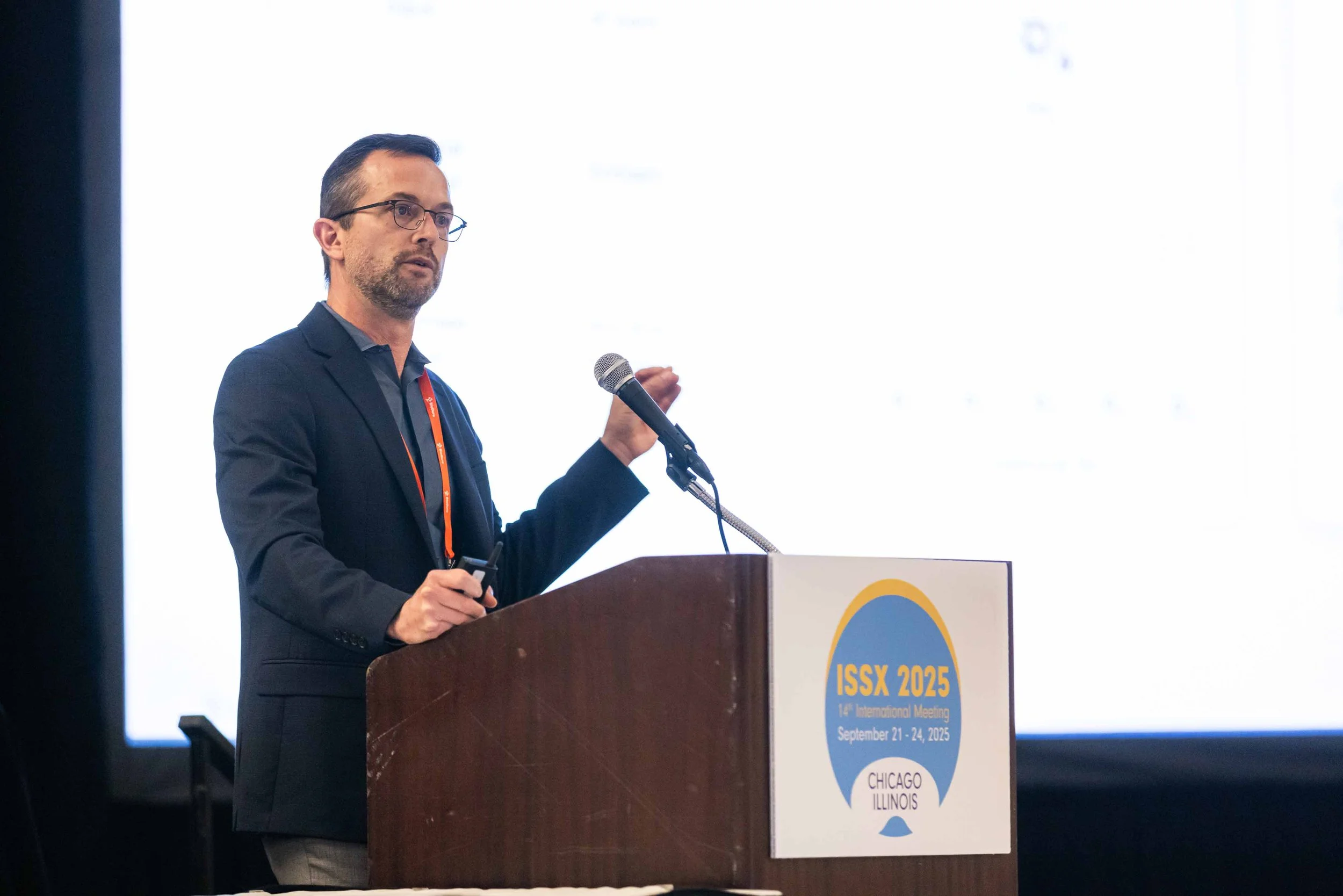 A man in a dark suit and glasses giving a presentation at a conference with a microphone and a remote clicker, standing behind a podium with a sign that reads ISSX 2025, 14th International Meeting, September 21-24, 2025, Chicago Illinois.