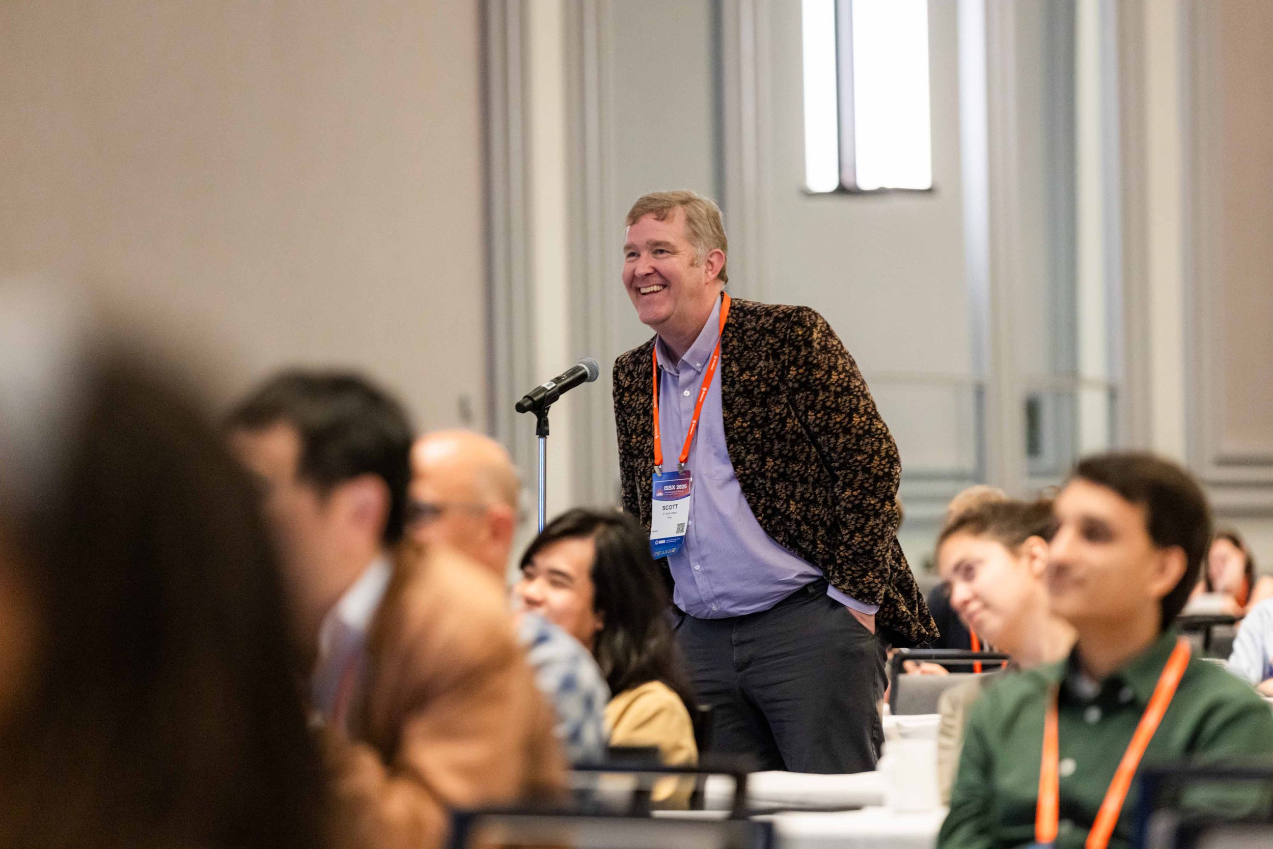 A man with a patterned blazer and light shirt is smiling and standing at a microphone during a conference, with other seated attendees around him.