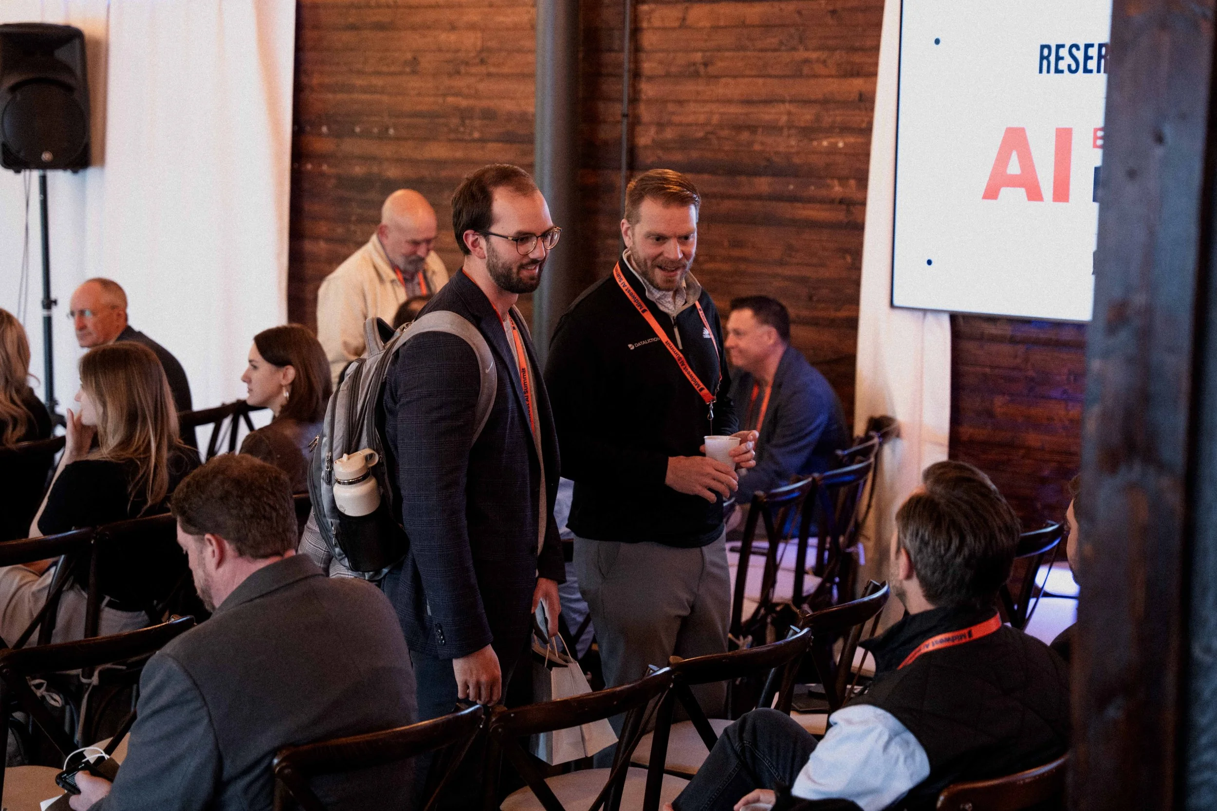 Two men with conference badges talking at a professional event, seated among others in a room with wooden walls and a large screen displaying text.