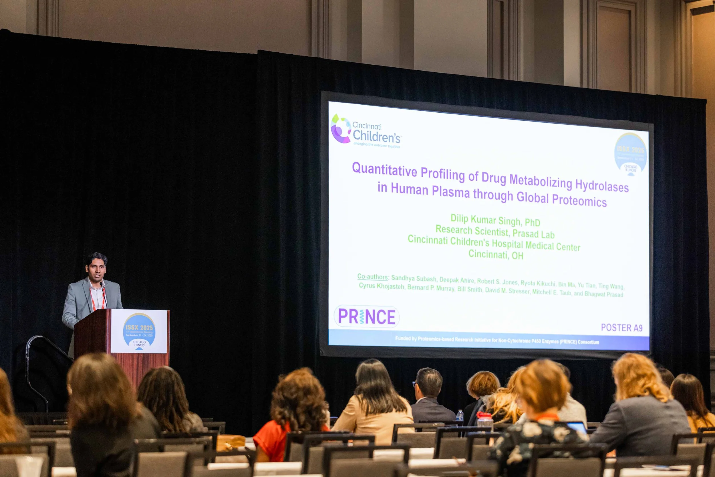 A man presenting a scientific poster at a conference, with audience members seated in front of him and a large screen displaying the title of the presentation.