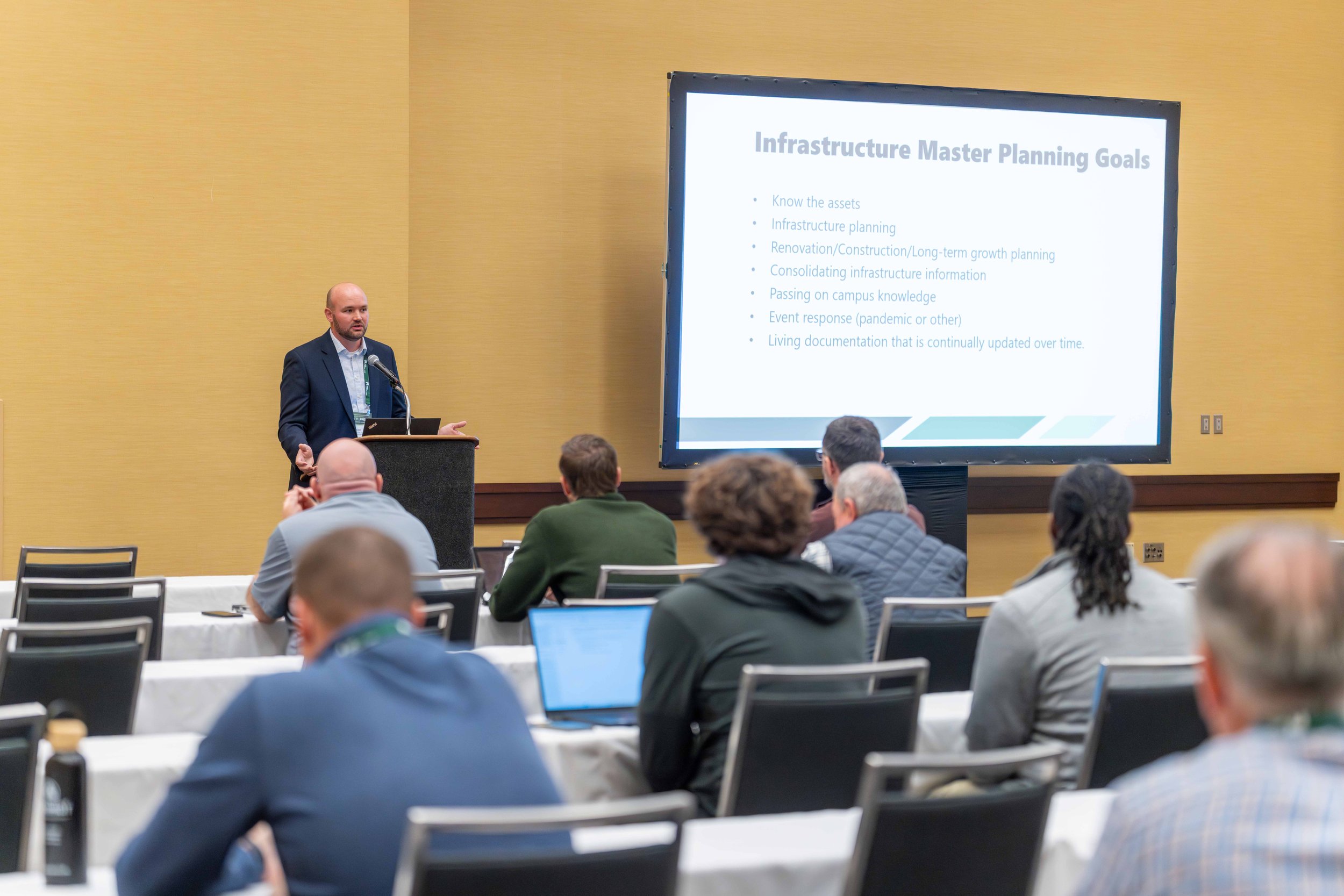 A man in a dark suit giving a presentation at a conference, standing behind a podium with a microphone, while a large screen displays the slide titled 'Infrastructure Master Planning Goals'. Several people are seated in the audience, some taking note
