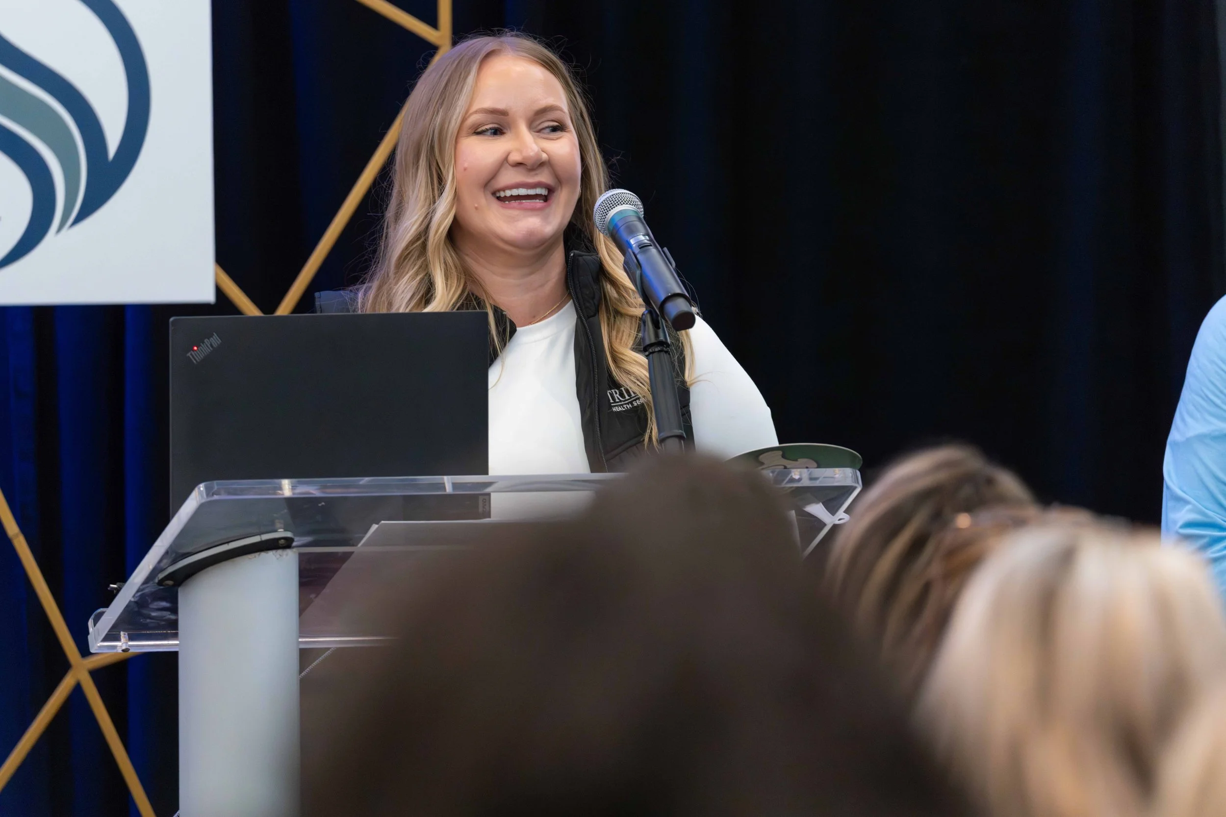 A woman with long blonde hair smiling and speaking at a podium with a microphone during a presentation or conference.