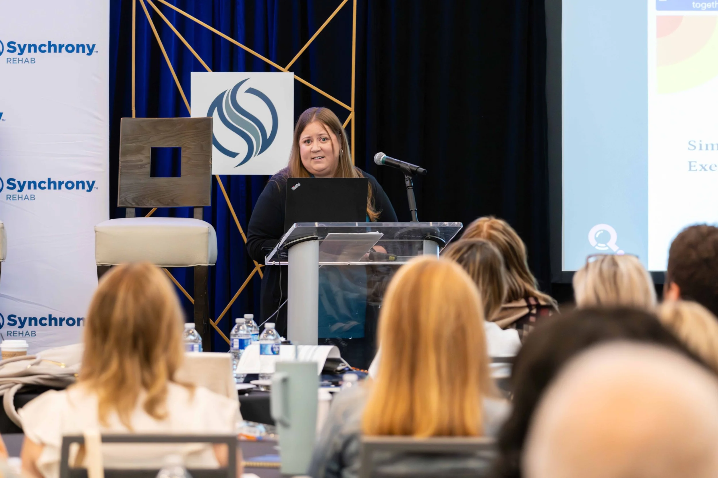 A woman giving a presentation at a conference, standing behind a podium with a laptop, in front of an audience, with a backdrop displaying the 'Synchrony REHAB' logo and a large screen with a slide.