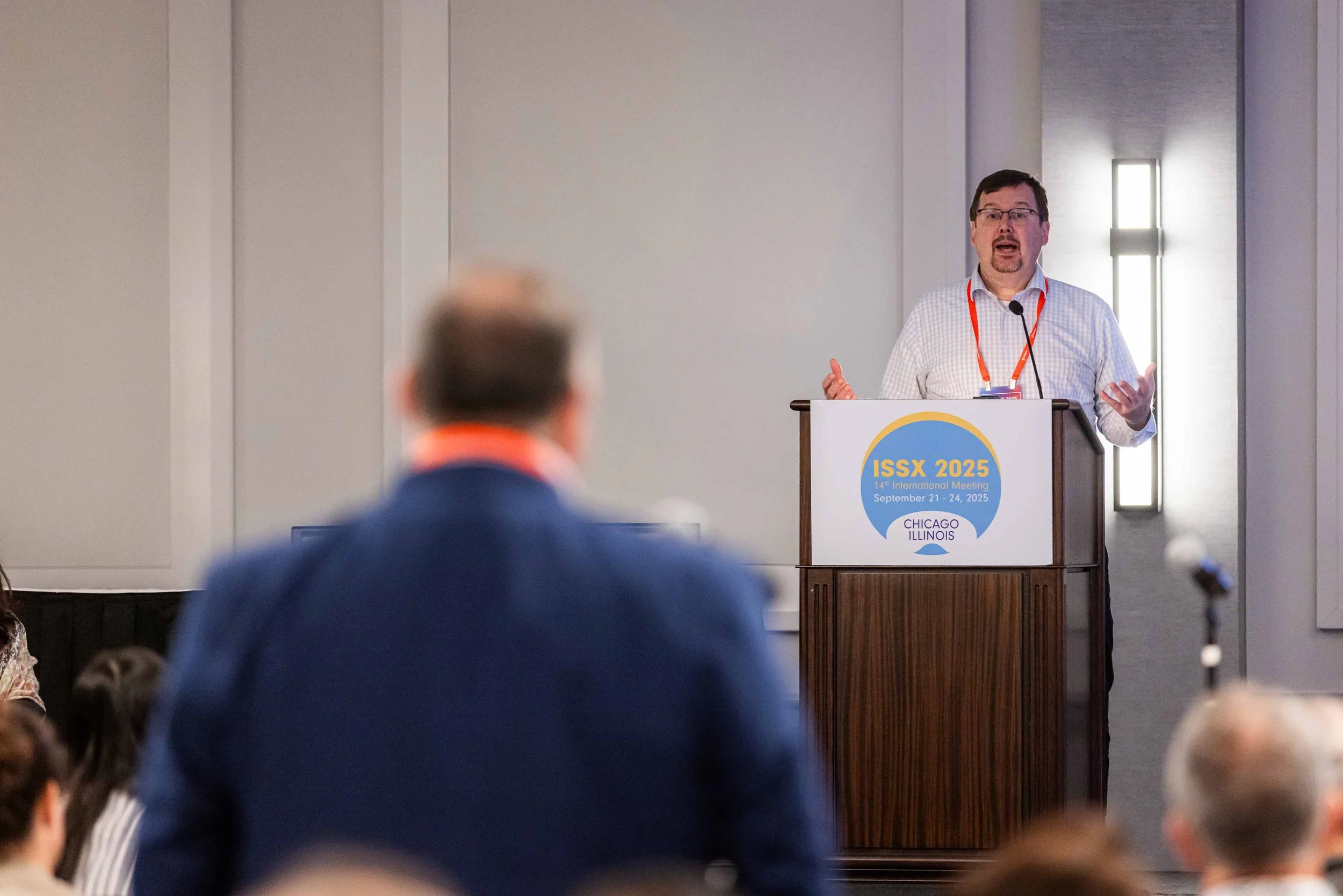 A man speaking at a conference podium with a sign that reads 'ISSX 2025, 14th International Meeting, September 21-24, 2025, Chicago Illinois.' The audience is visible, with a person in a blue jacket in the foreground.