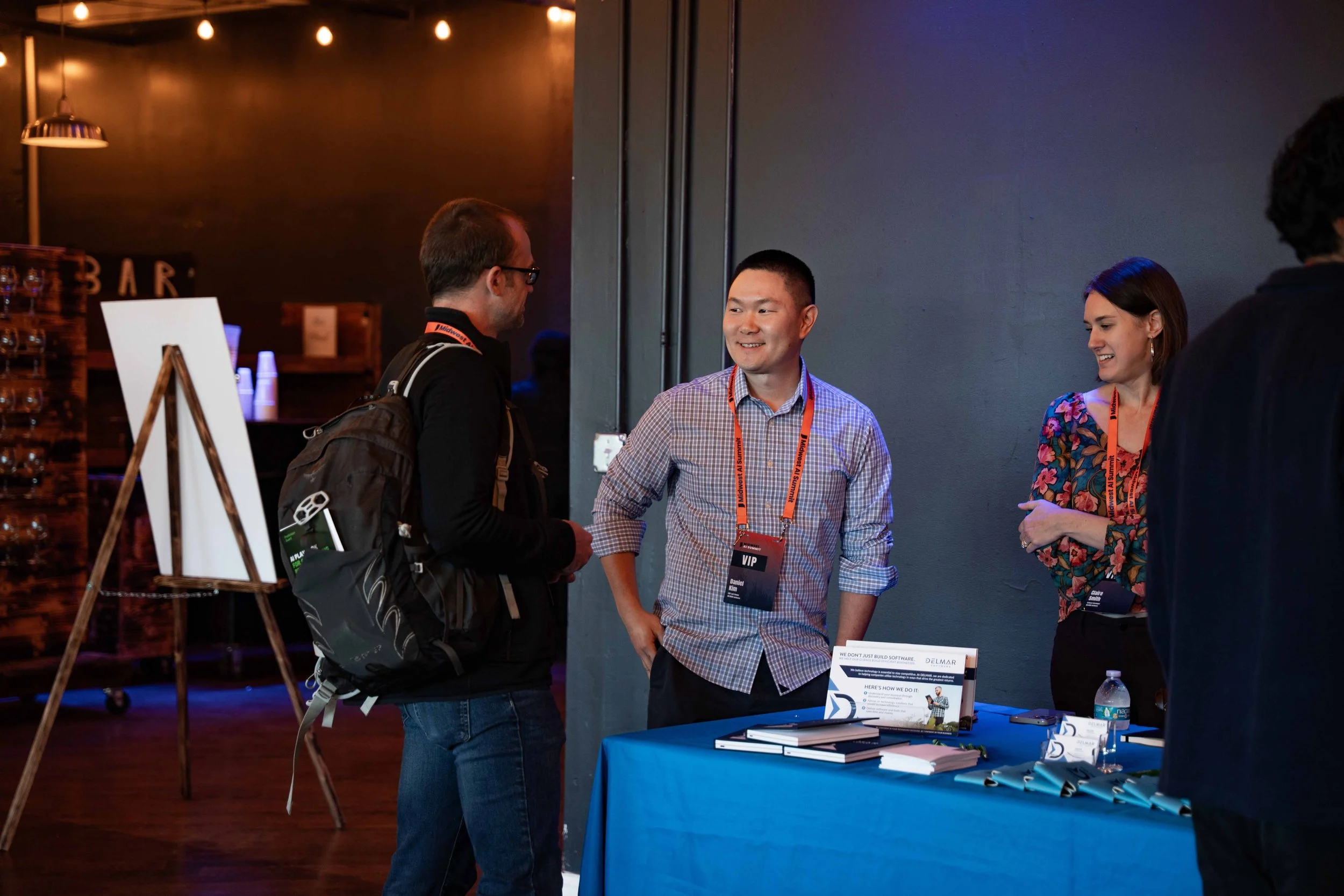 Three people are engaged in conversation at a conference registration table. One man with glasses and a backpack is talking to a smiling man in a checkered shirt with a VIP badge. A woman is standing behind the table, smiling, with a water bottle in 