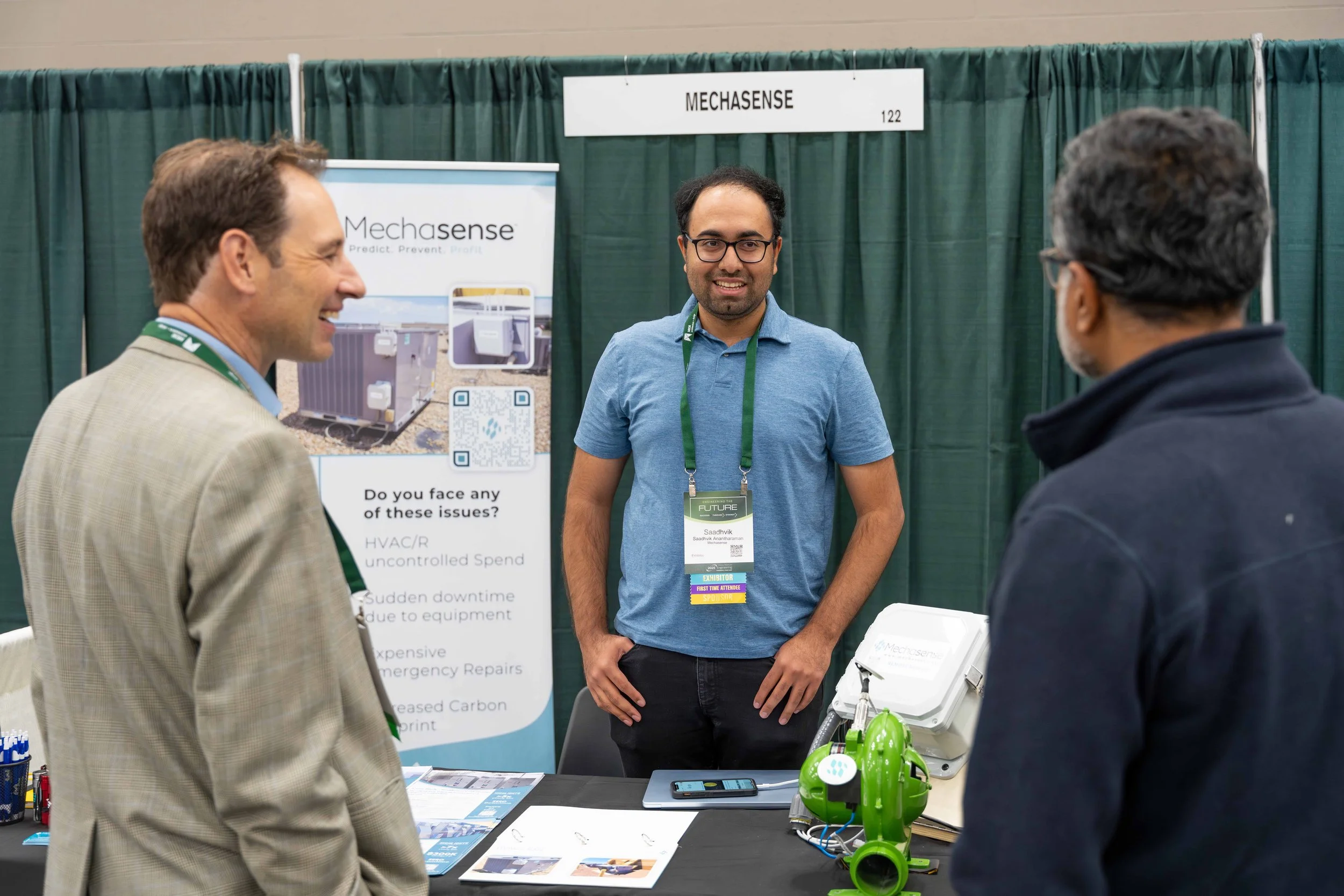 Three men are standing at a trade show booth with green curtains and a 'Mechasense' sign, engaging in conversation. The booth has informational materials, a green mask, and a white device.