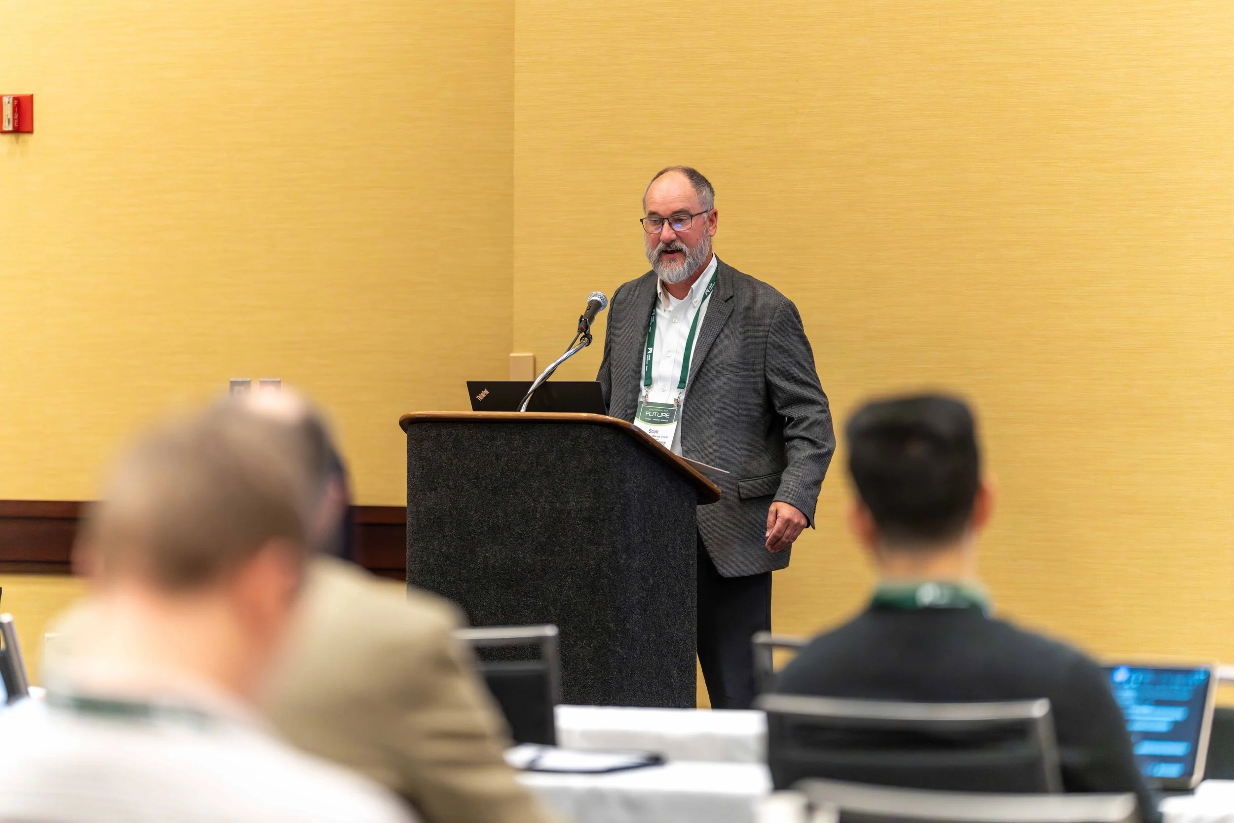 A man with glasses and a beard standing at a podium giving a presentation in a conference room, wearing a gray blazer and white shirt, with a laptop on the podium and an audience seated in front.