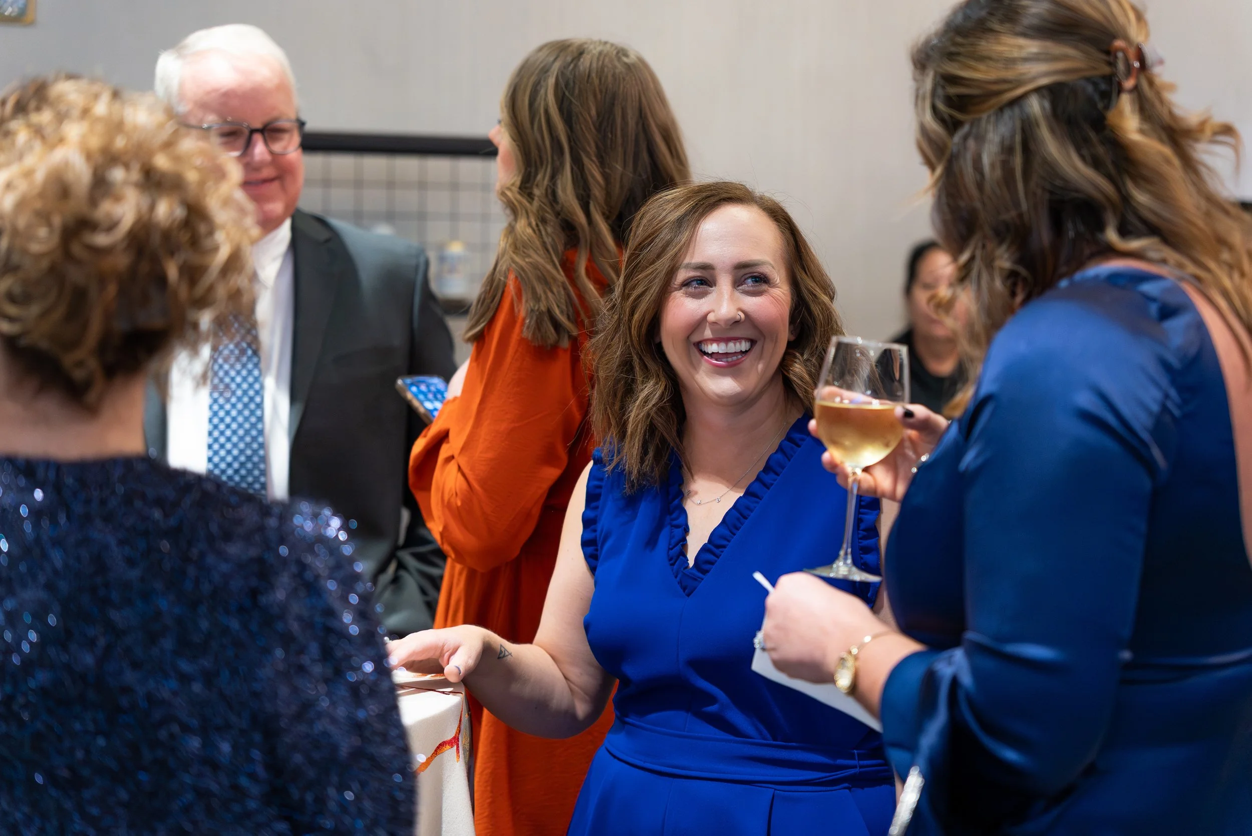 Group of women at a social event, one woman in a blue dress smiling and holding a glass of white wine, others engaging in conversation.