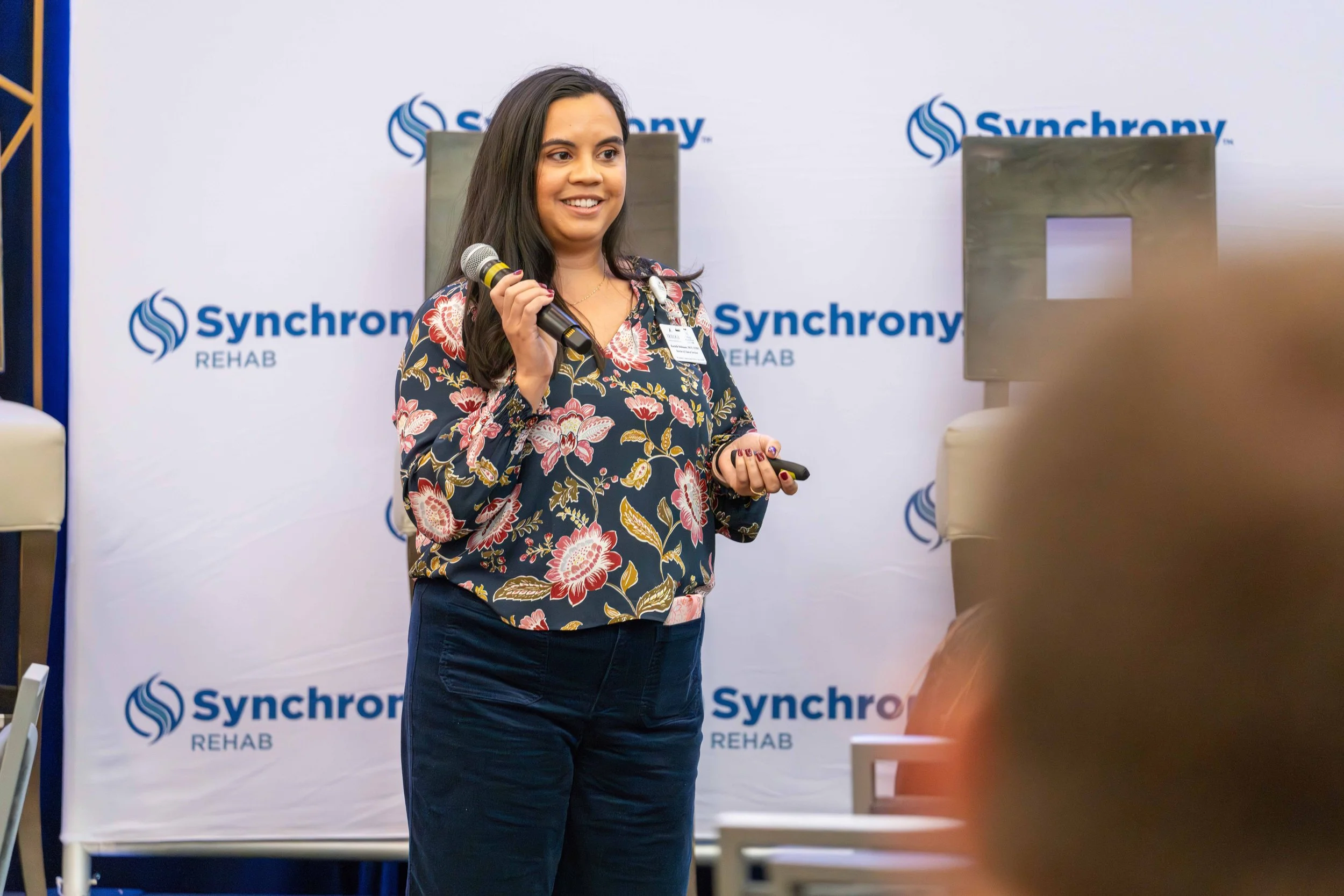 Woman holding microphone and presenting at Synchrony Rehab event