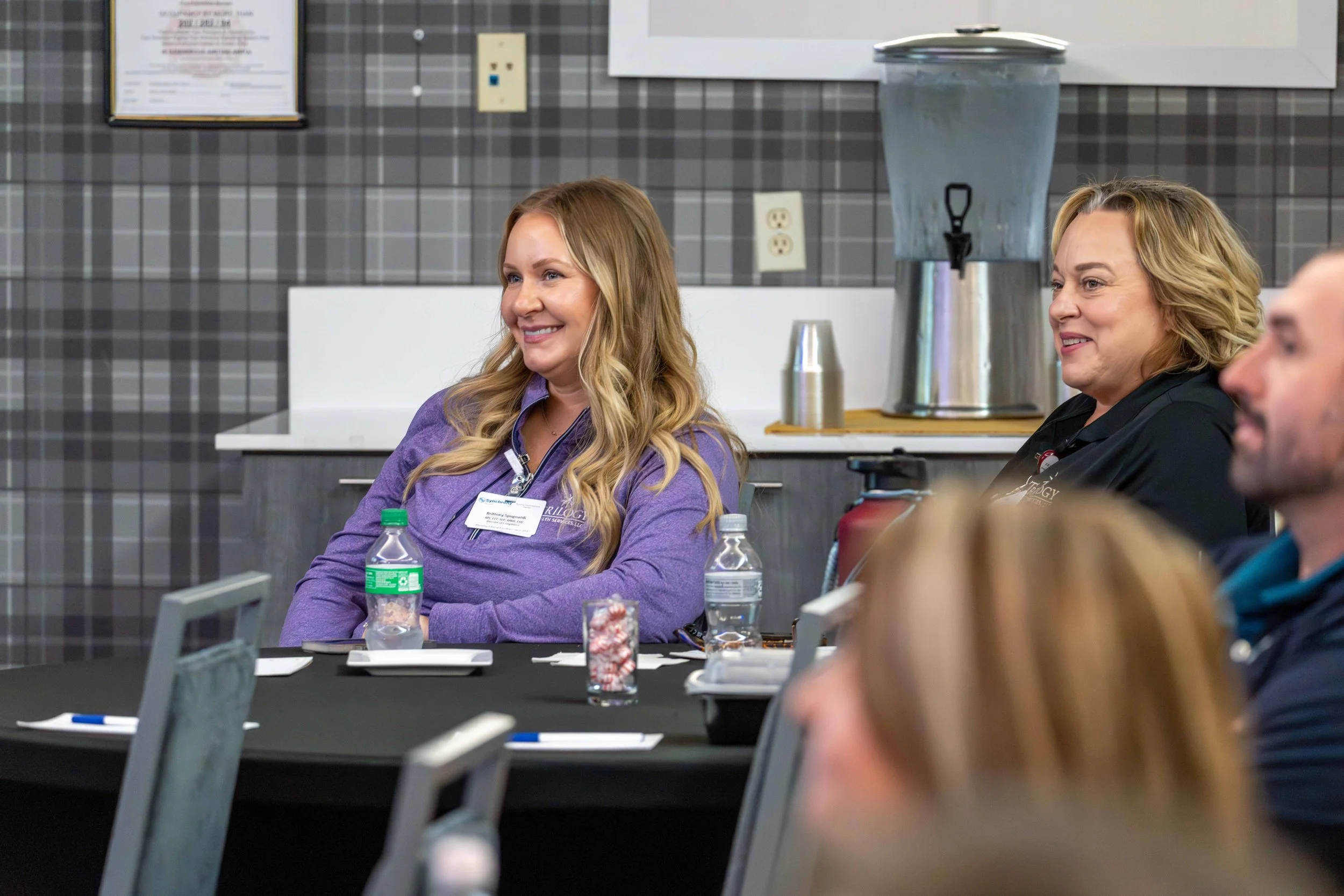 Group of people sitting at a conference table in a meeting room, listening and smiling. Two women are prominently visible, one in a purple shirt with a name badge and the other in black, with drinks and notepads on the table.