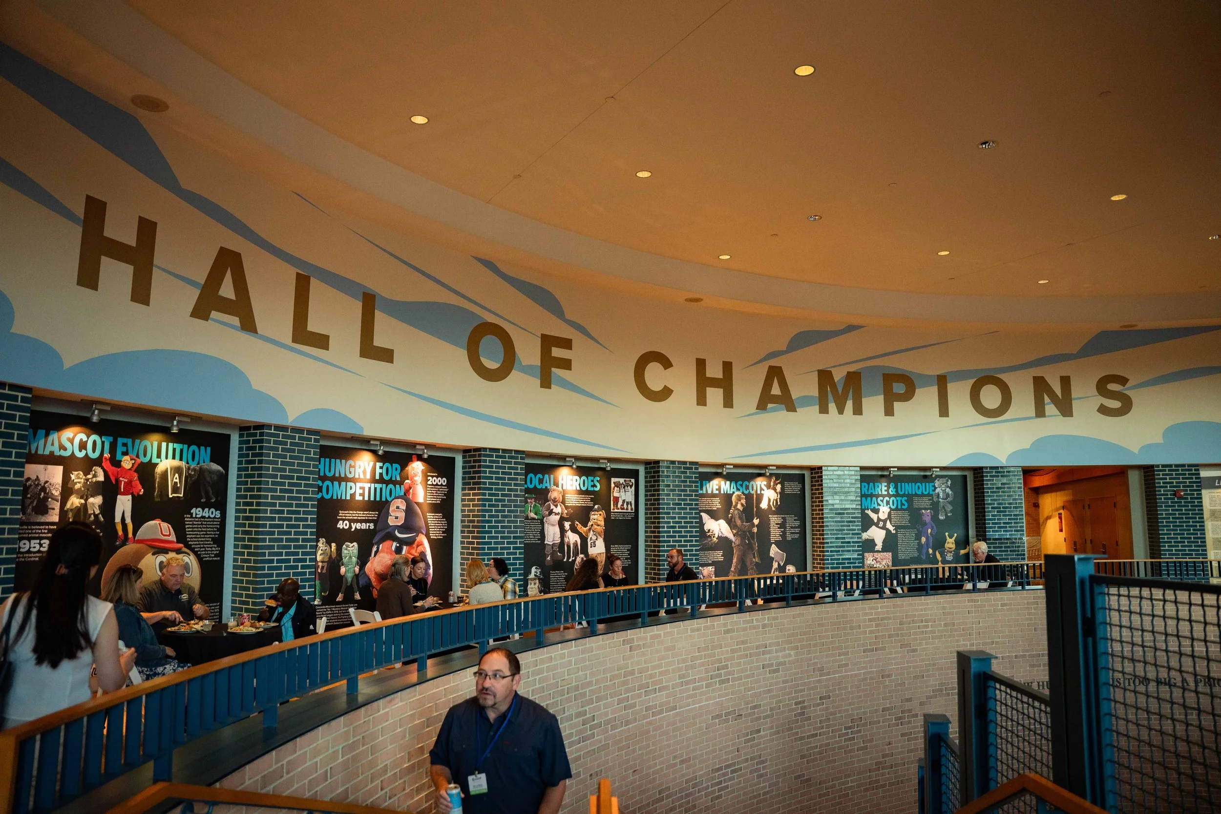 Indoor exhibit area titled 'Hall of Champions' with display panels and people walking along a curved balcony railing.
