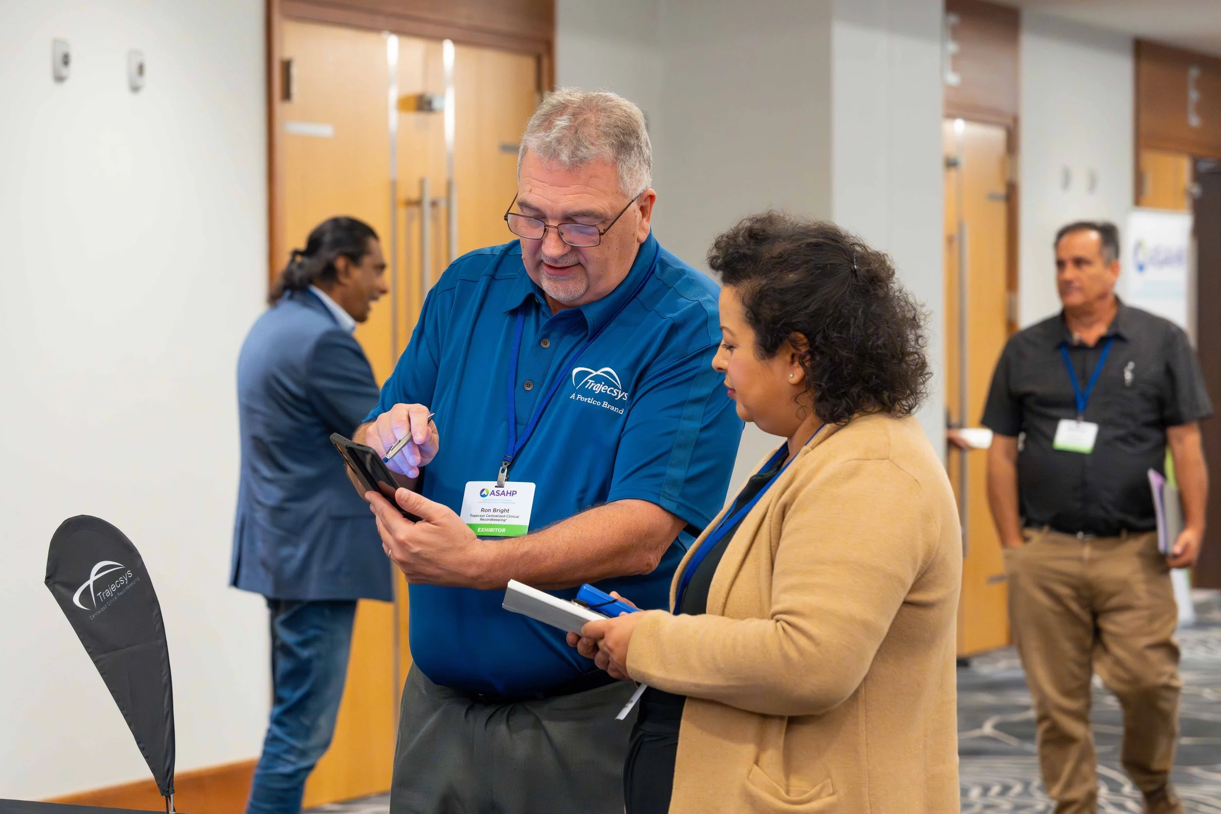 Two people are standing and looking at a smartphone together at a conference. One man is wearing a blue shirt with a badge and the other woman is wearing a tan blazer with a notepad. In the background, there are two other men, one in a black shirt an
